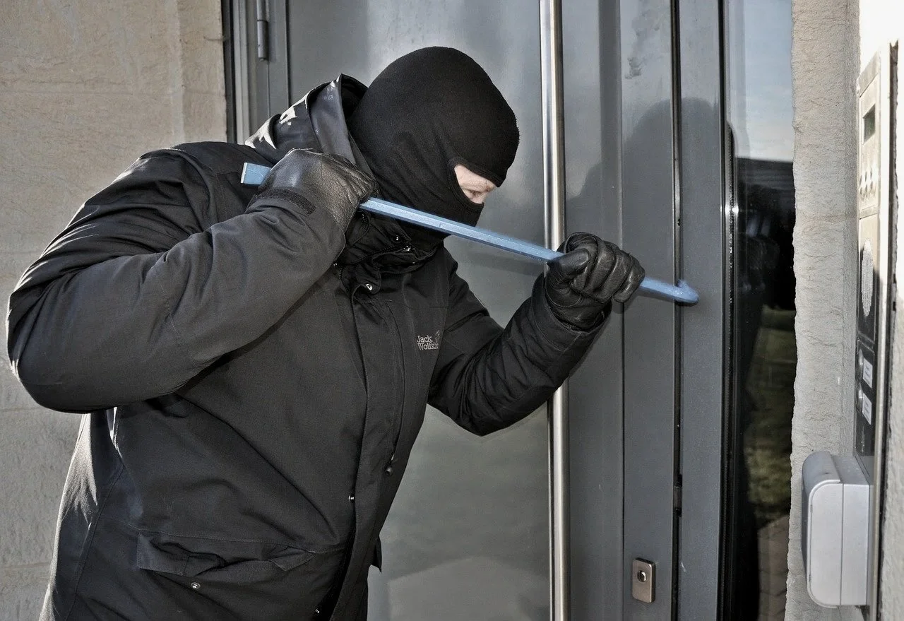 Ein maskierter Einbrecher mit schwarzer Jacke und Handschuhen versucht, eine Tür mit einem Hebel aufzubrechen.