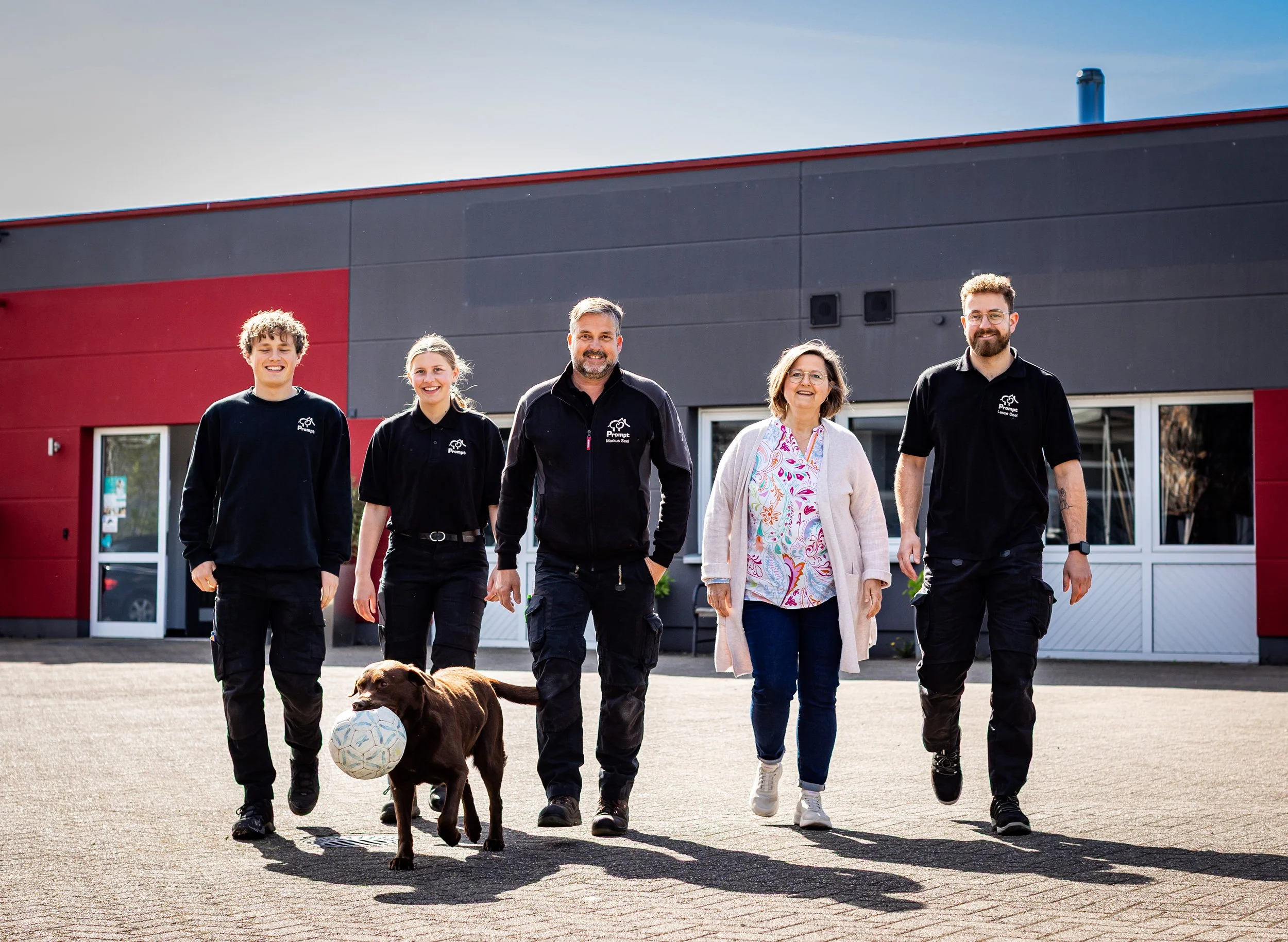 Die Belegschaft der Firma prompt läuft lachend vor dem Gebäude der Firma entlang. Der Firmenhund hat einen Ball im Maul und läuft vorweg.