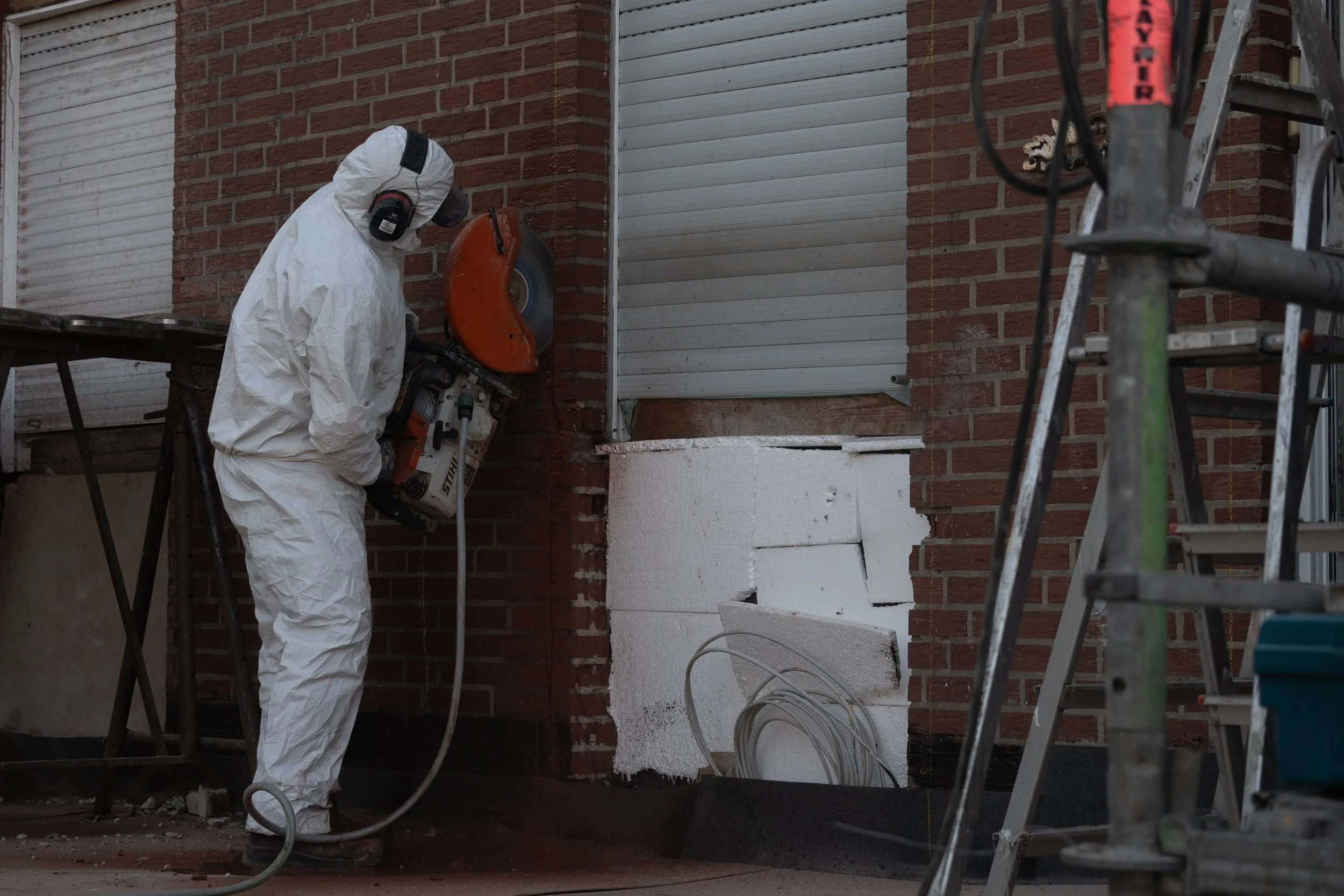 Bauarbeiter in Schutzkleidung arbeitet an einem Haus mit Ziegelwänden und Isolierung. Es stehen Leitern im Bild, und die Arbeiter verwendet einen Motortrennschleifer von Stihl.  
