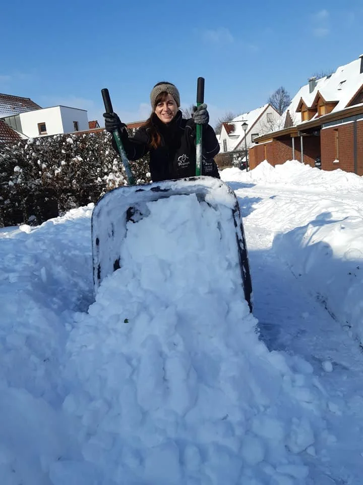 Frau mit Mütze und Handschuhen schaufelt Schnee vor einem Haus im Winter.
