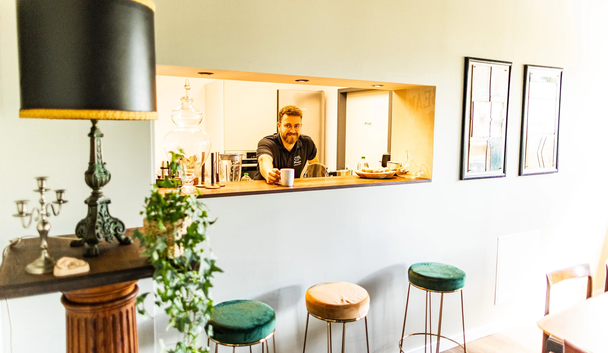 Ein Mann steht an einem Küchen- oder Bartresen, hält eine Tasse und lächelt. Im Vordergrund sind dekorative Gegenstände wie Kerzen, ein Glas-Obstbehälter und Pflanzen, im Hintergrund sind Bilder an der Wand. Es herrscht ein gemütliches Ambiente.