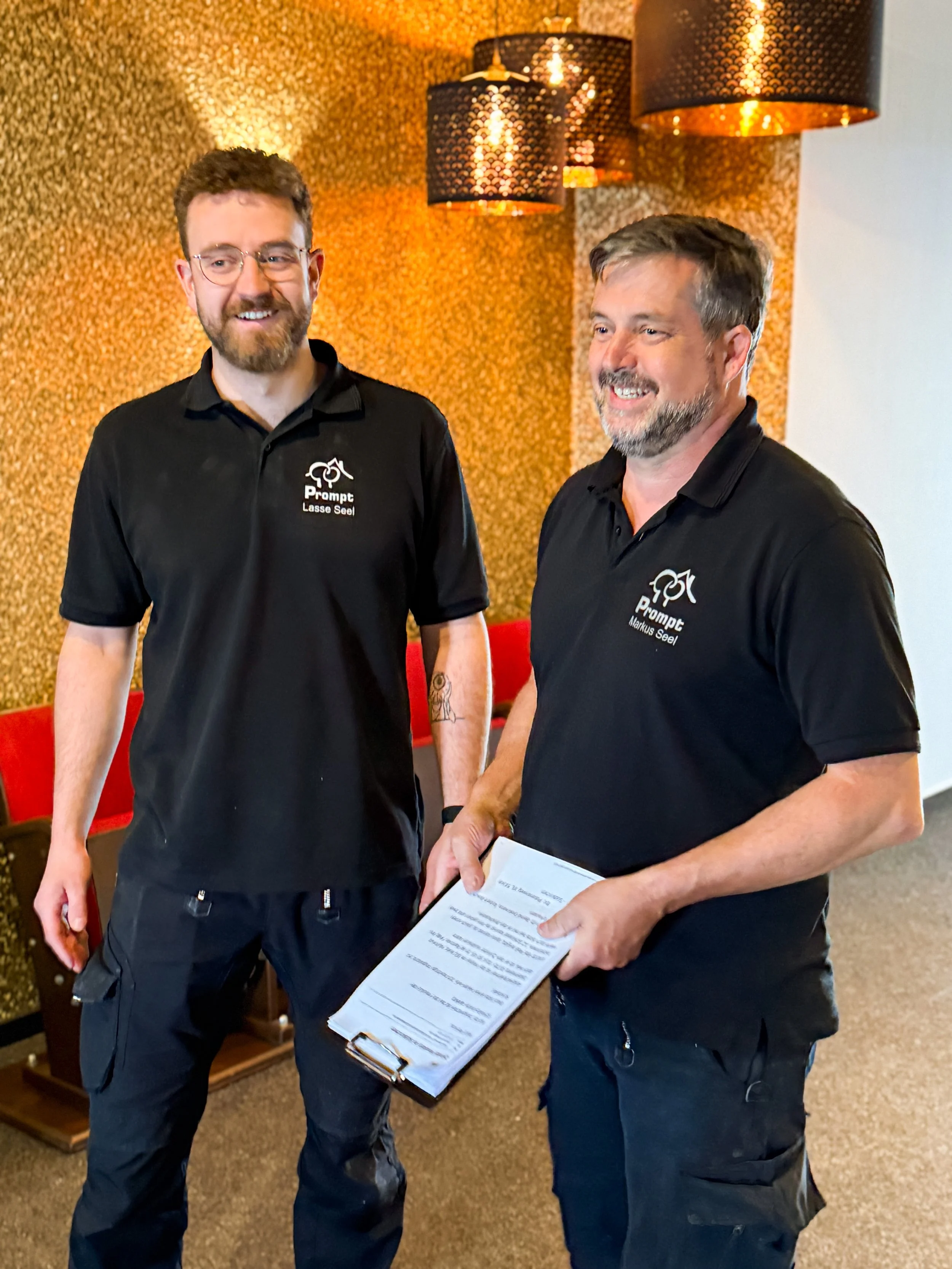 Zwei Männer in schwarzen Polo-Shirts mit Firmenlogo stehen in einem Raum mit goldener Wand und schwarzen Lampen, während einer eine Palette mit Dokumenten hält.