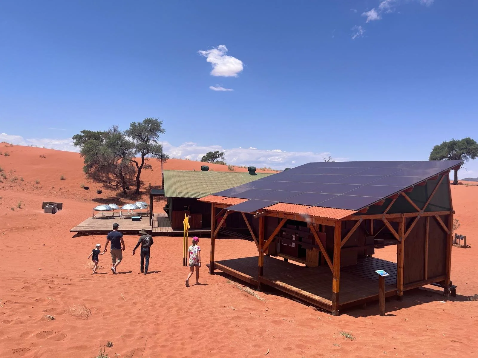 A family of five walking towards a small wooden house with solar panels in a desert landscape with red sand, sparse trees, and a bright blue sky.
