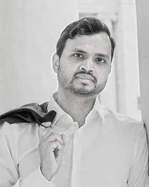 Black and white portrait of a man with facial hair, wearing a shirt and holding a coat over his shoulder.