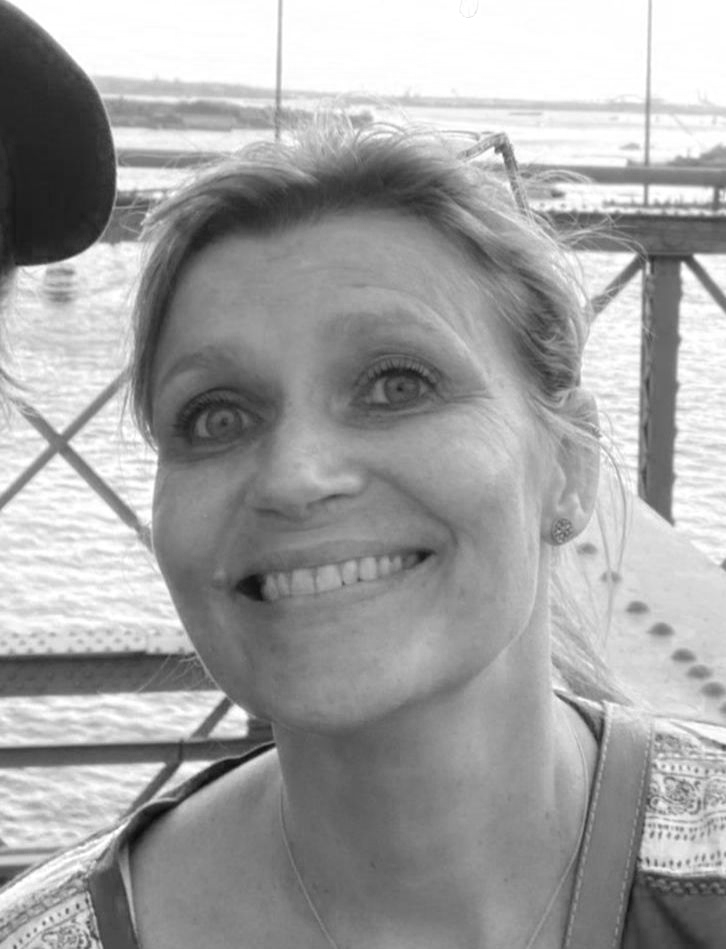 A woman smiling outdoors near a body of water with a bridge or dock structure in the background.