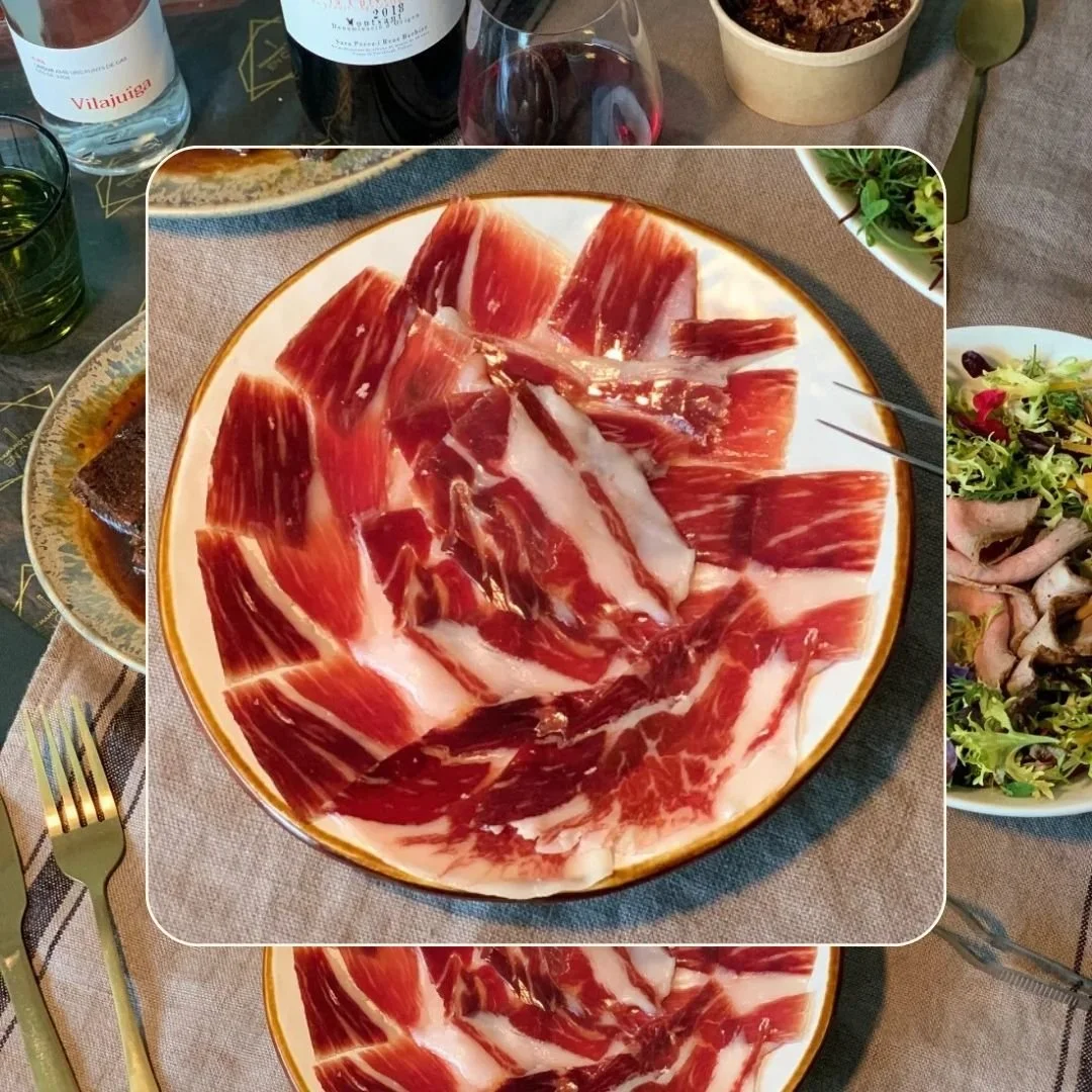 Plate of thinly sliced cured Iberic ham, on a dining table with wine bottles, glasses, and other dishes.