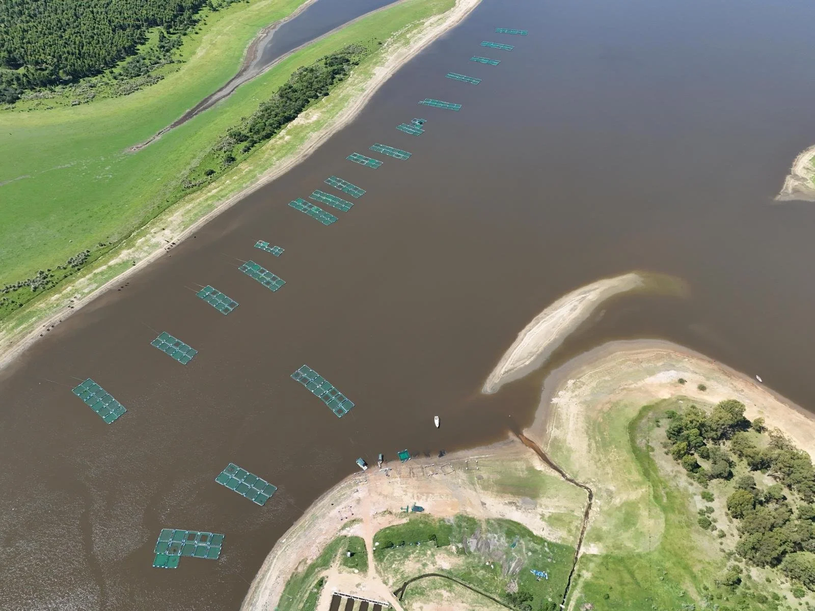 Aerial view of a body of water with floating platforms, a boat near the platforms, and green land with trees and sandy shores on both sides.