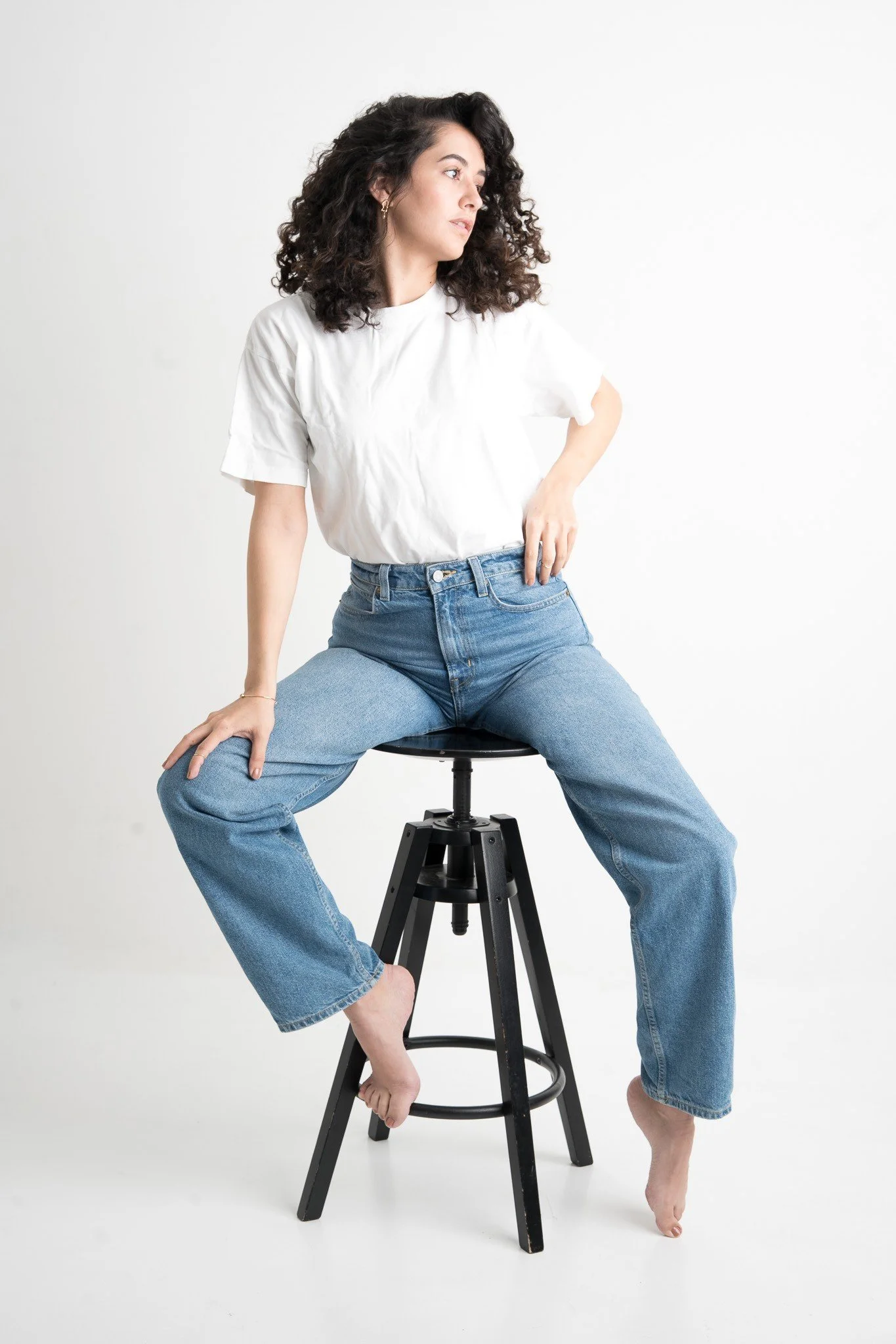 Frau sitzt nackt auf Hocker, trägt Jeans und T-Shirt, Blick seitlich, minimalistischer weißer Hintergrund.