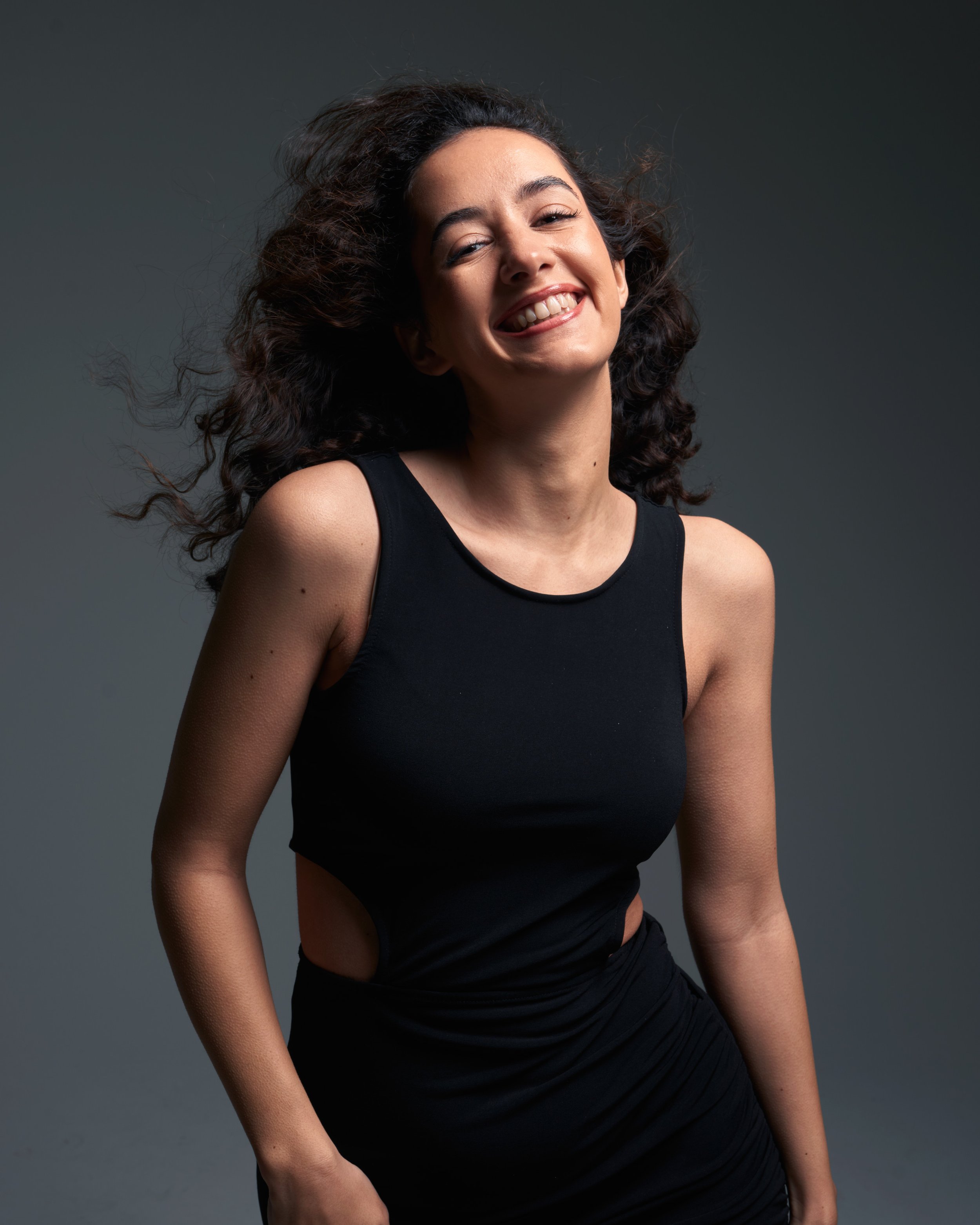 Eine lächelnde junge Frau mit dunklen, lockigen Haaren trägt ein schwarzes ärmelloses Oberteil und ein schwarzes Kleidungstück.