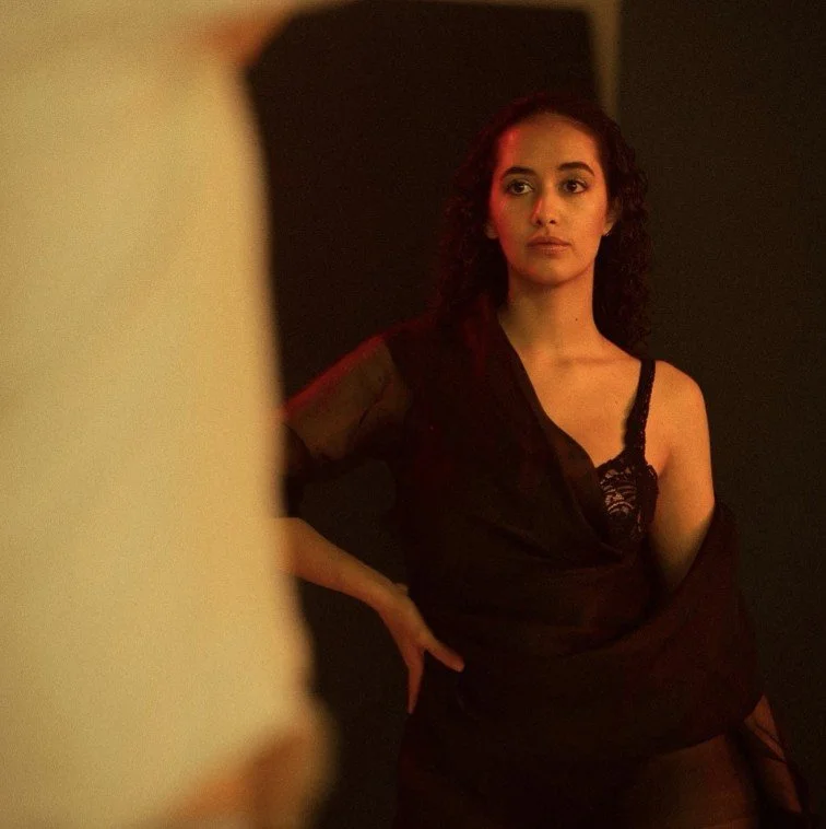 Junge Frau mit Locken trägt schwarzes Top mit Spitze, steht vor dunklem Hintergrund, blickt nach rechts, hat eine Hand an der Hüfte.