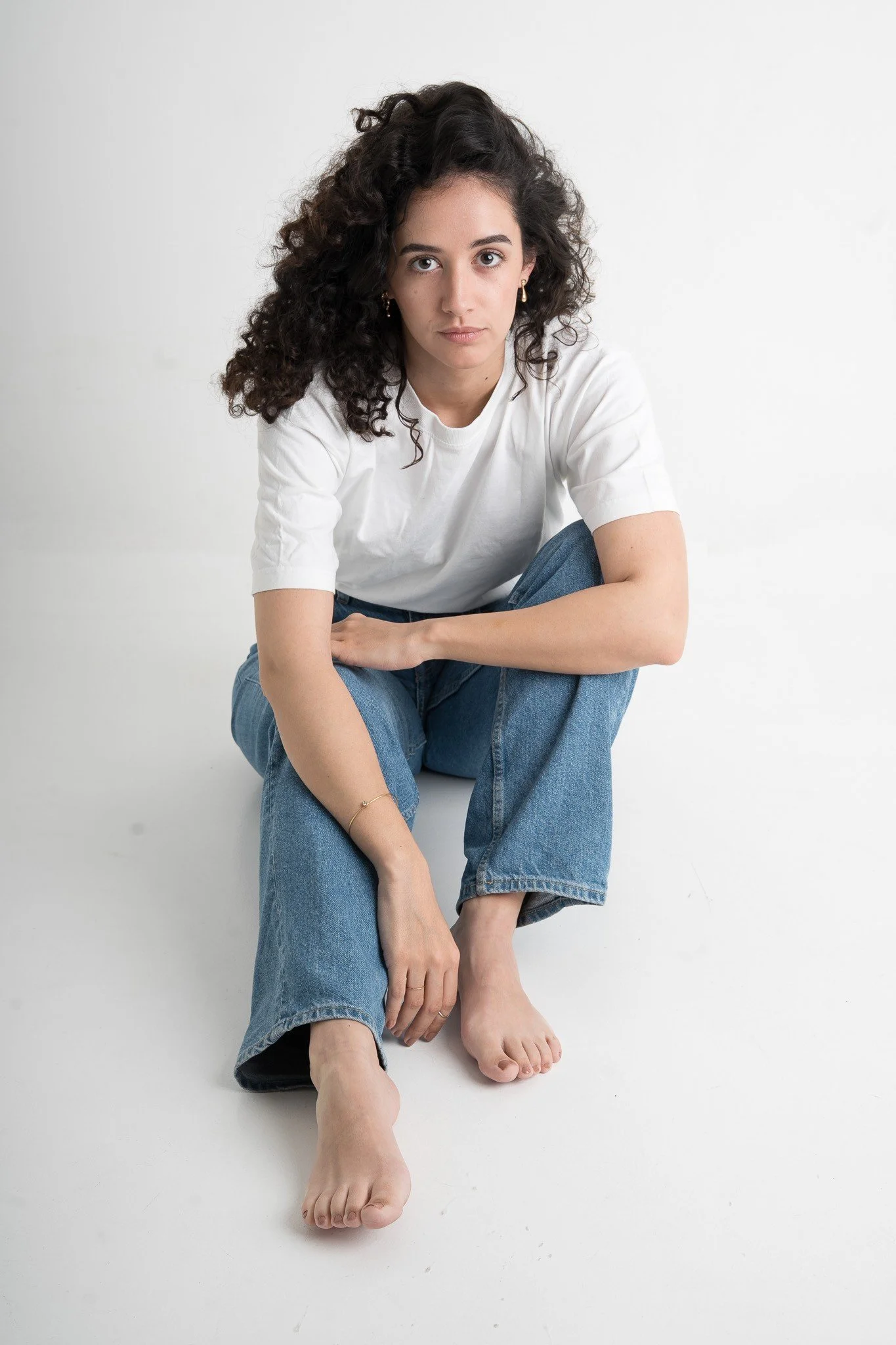 Junge Frau mit dunklen, lockigen Haaren, weißem T-Shirt und blauen Jeans, sitzt barfuß auf dem Boden vor neutralem, weißem Hintergrund.