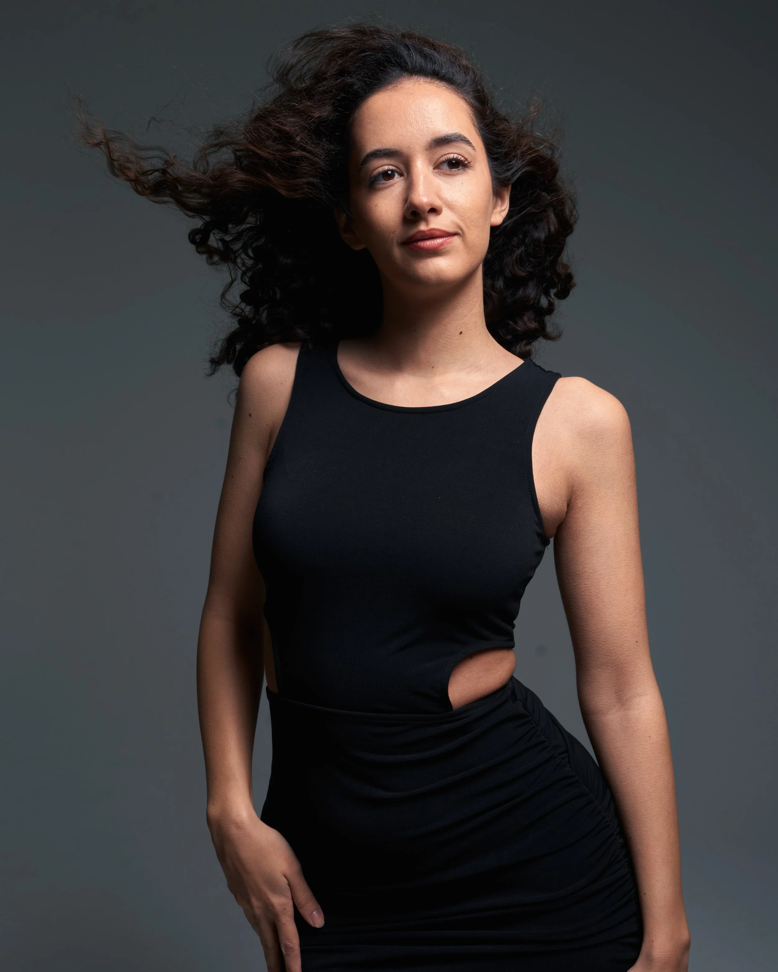 Junge Frau mit schwarzen, lockigen Haaren in einem schwarzen Kleid, steht vor grauem Hintergrund und blickt selbstbewusst in die Kamera.