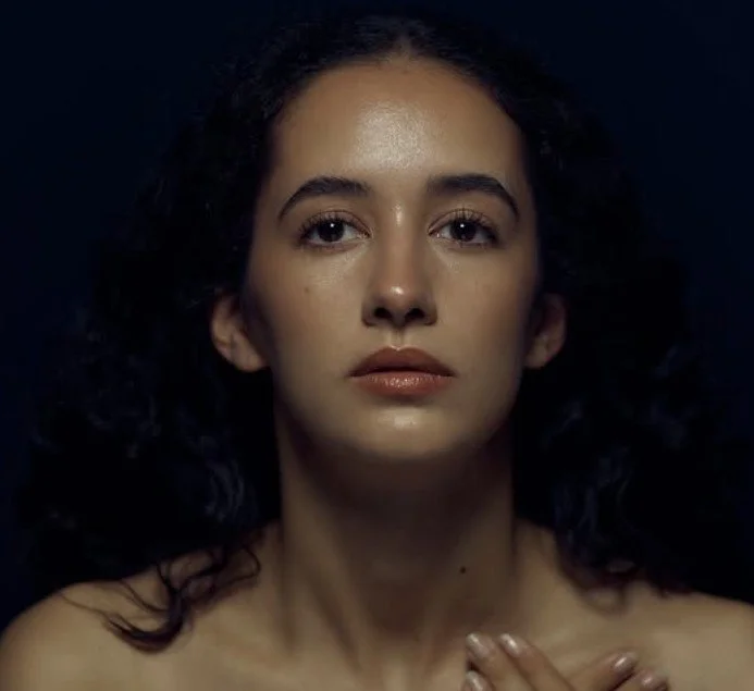 Nahaufnahme eines jungen Mannes mit dunklen, lockigen Haaren und natürlichem Make-up vor schwarzem Hintergrund.