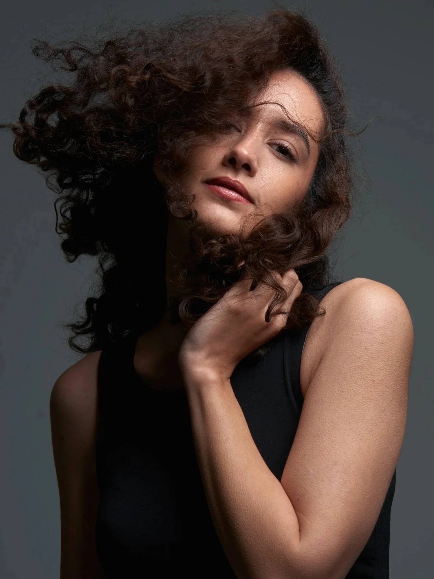 Eine junge Frau mit lockigem braunem Haar, die an ihrer Schulter greift, trägt ein schwarzes Top vor einem grauen Hintergrund.
