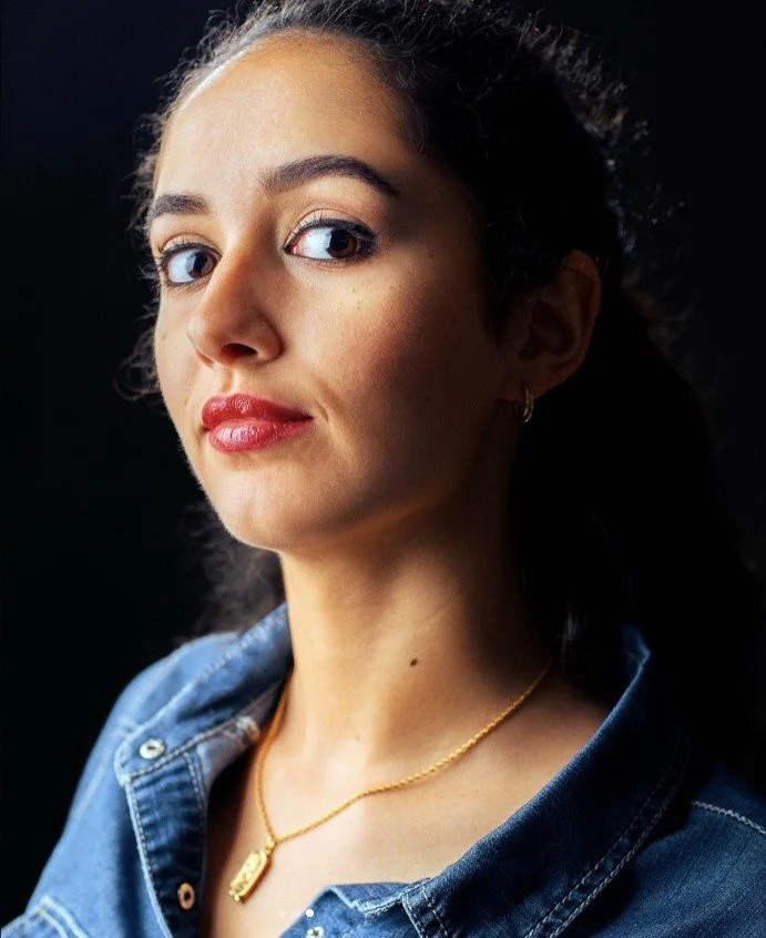 Nahaufnahme einer jungen Frau mit dunklem, gewelltem Haar, blauer Augen und rotem Lippenstift, trägt eine Jeansjacke und eine goldene Halskette vor schwarzem Hintergrund.