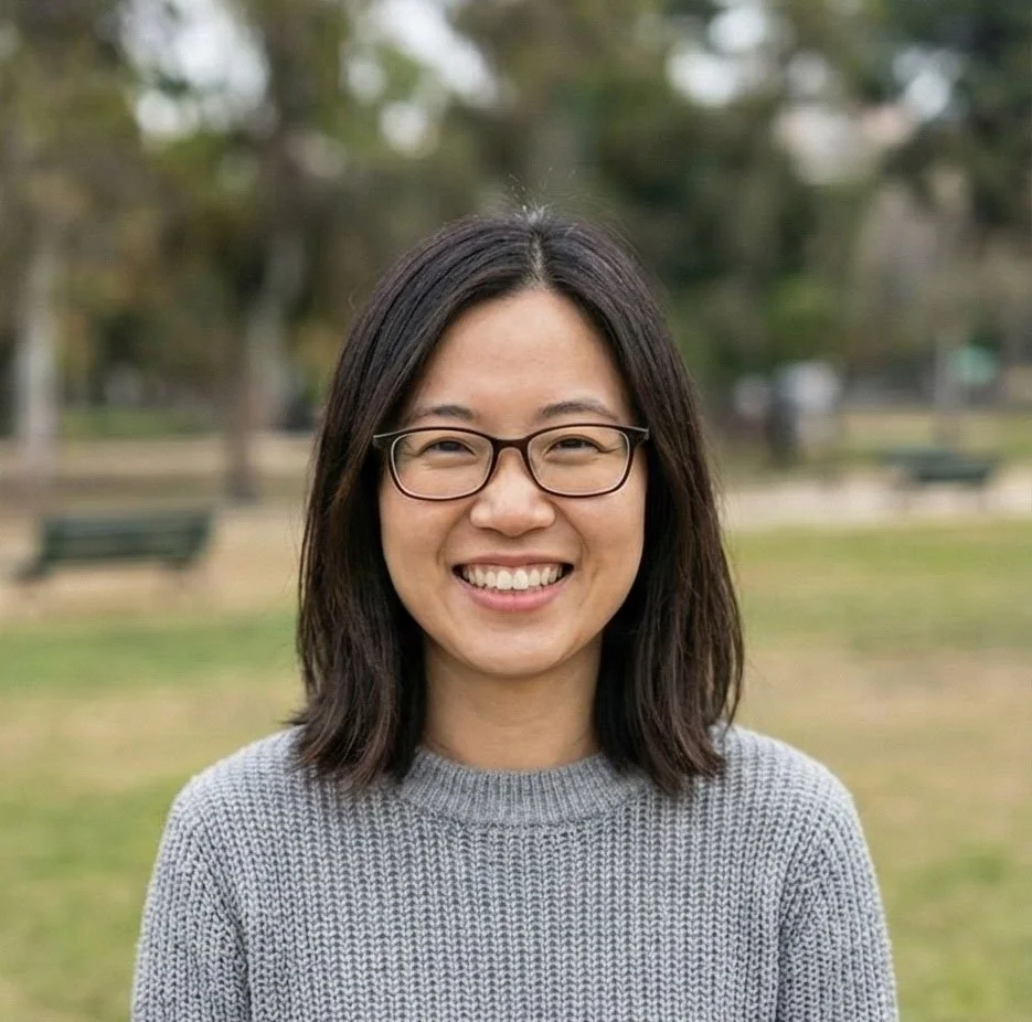 A woman with shoulder-length black hair, glasses, and a gray sweater standing outdoors in a park with trees and benches in the background, smiling at the camera.