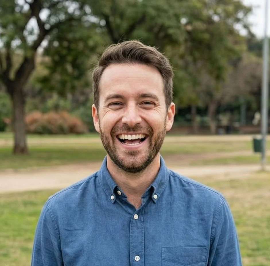 A smiling man with short brown hair and a beard, wearing a blue button-up shirt, standing outdoors on a grassy area with trees in the background.