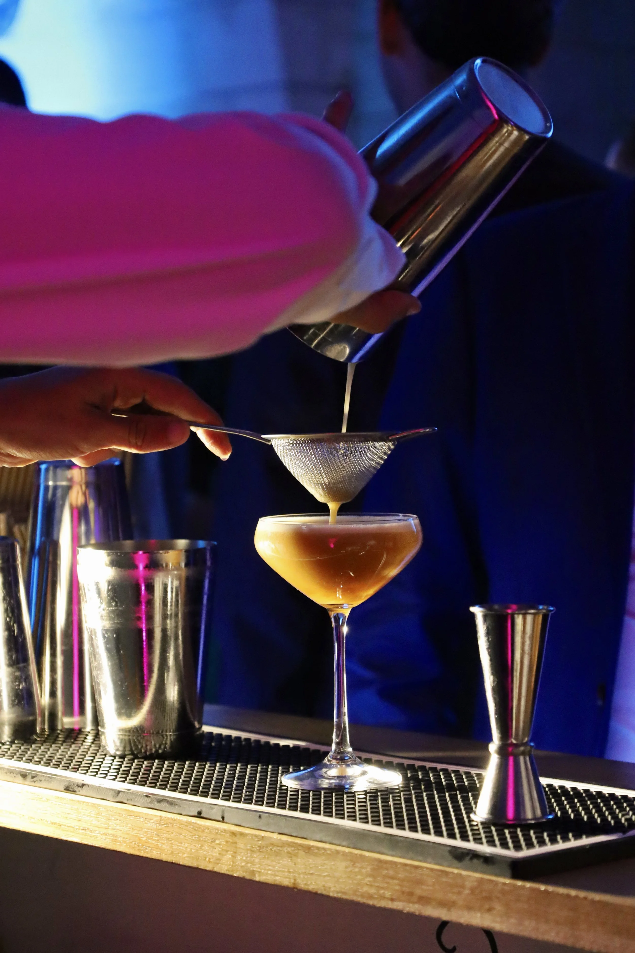 Une personne filtre un cocktail à l'aide d'une passoire fine dans un verre à cocktail, le tout sur un bar avec des outils de bartender en aluminium.