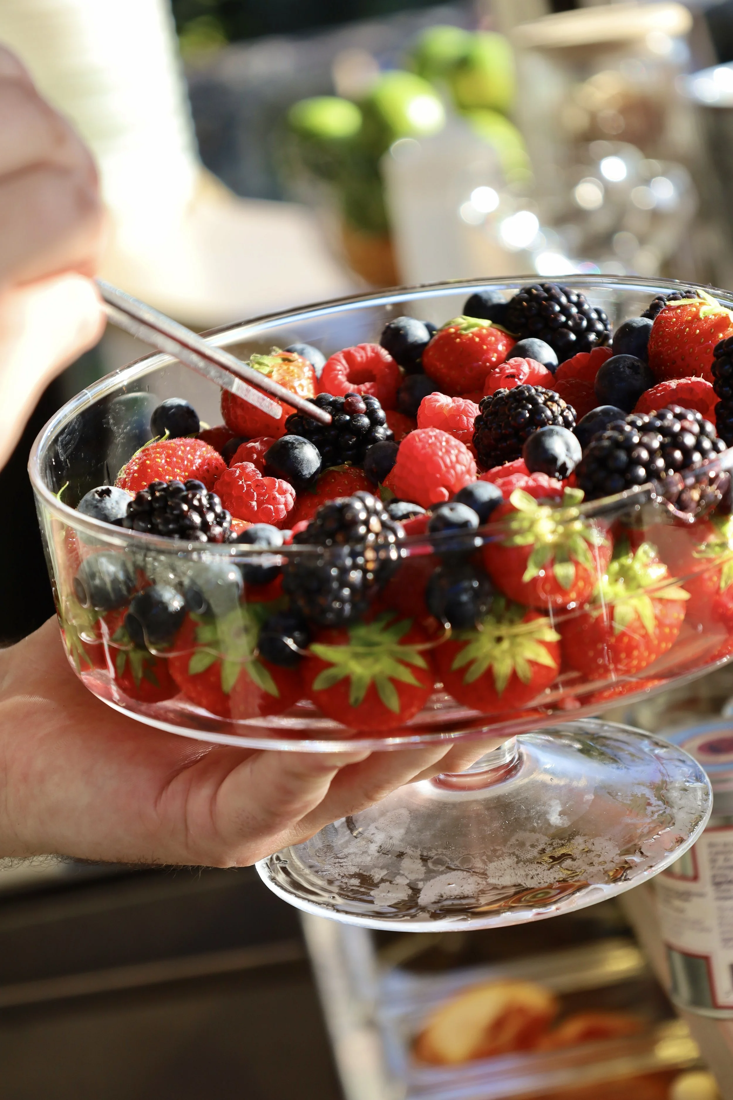 Une main tient un grand plat en verre rempli de fraises, framboises, mûres et myrtilles, avec une personne en arrière-plan qui semble goûter ou ajouter quelque chose.