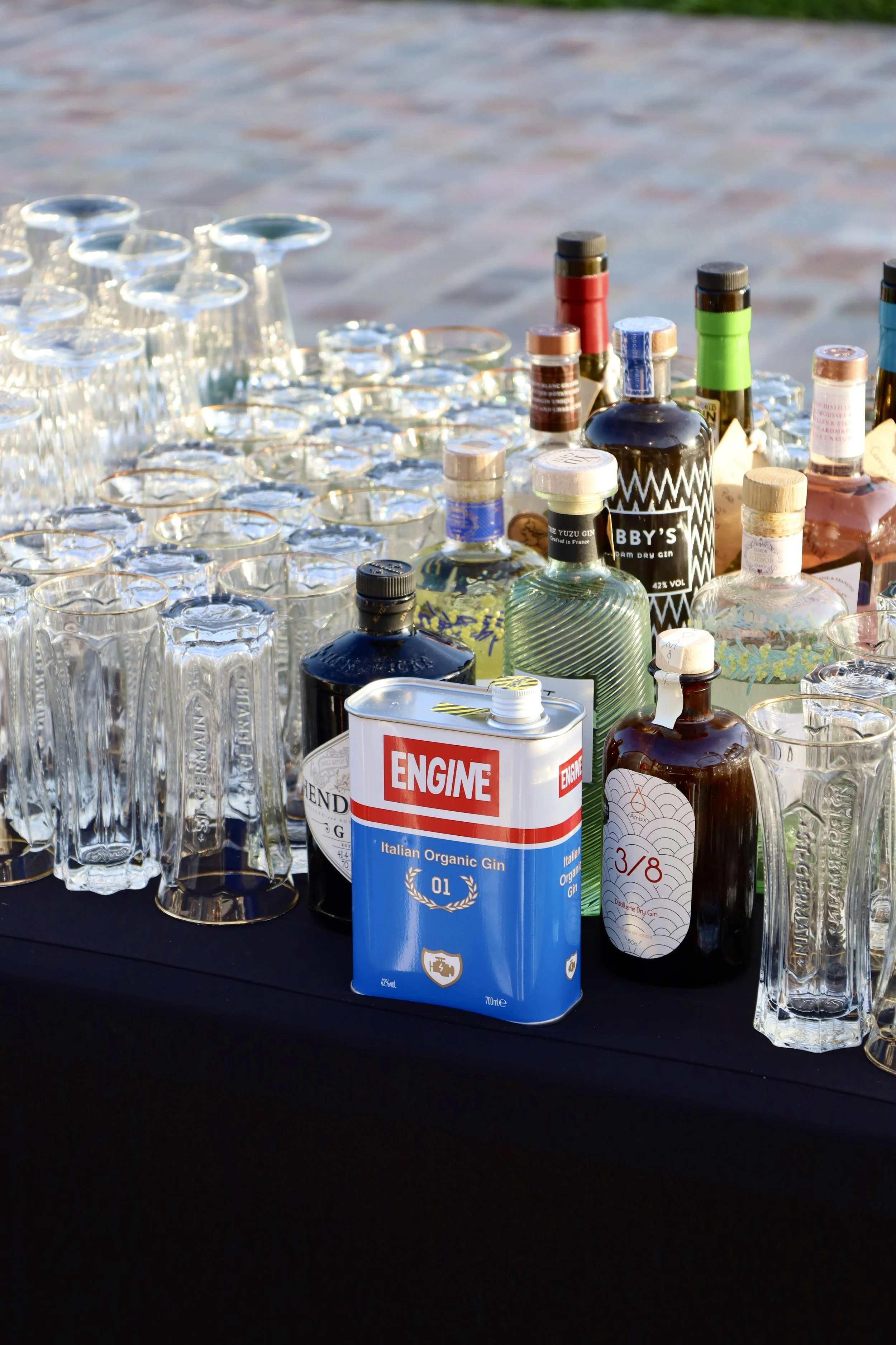Une variété de bouteilles d'alcool, de verres et une boîte d'gin organisés sur une table lors d'une fête ou d'un événement en extérieur.