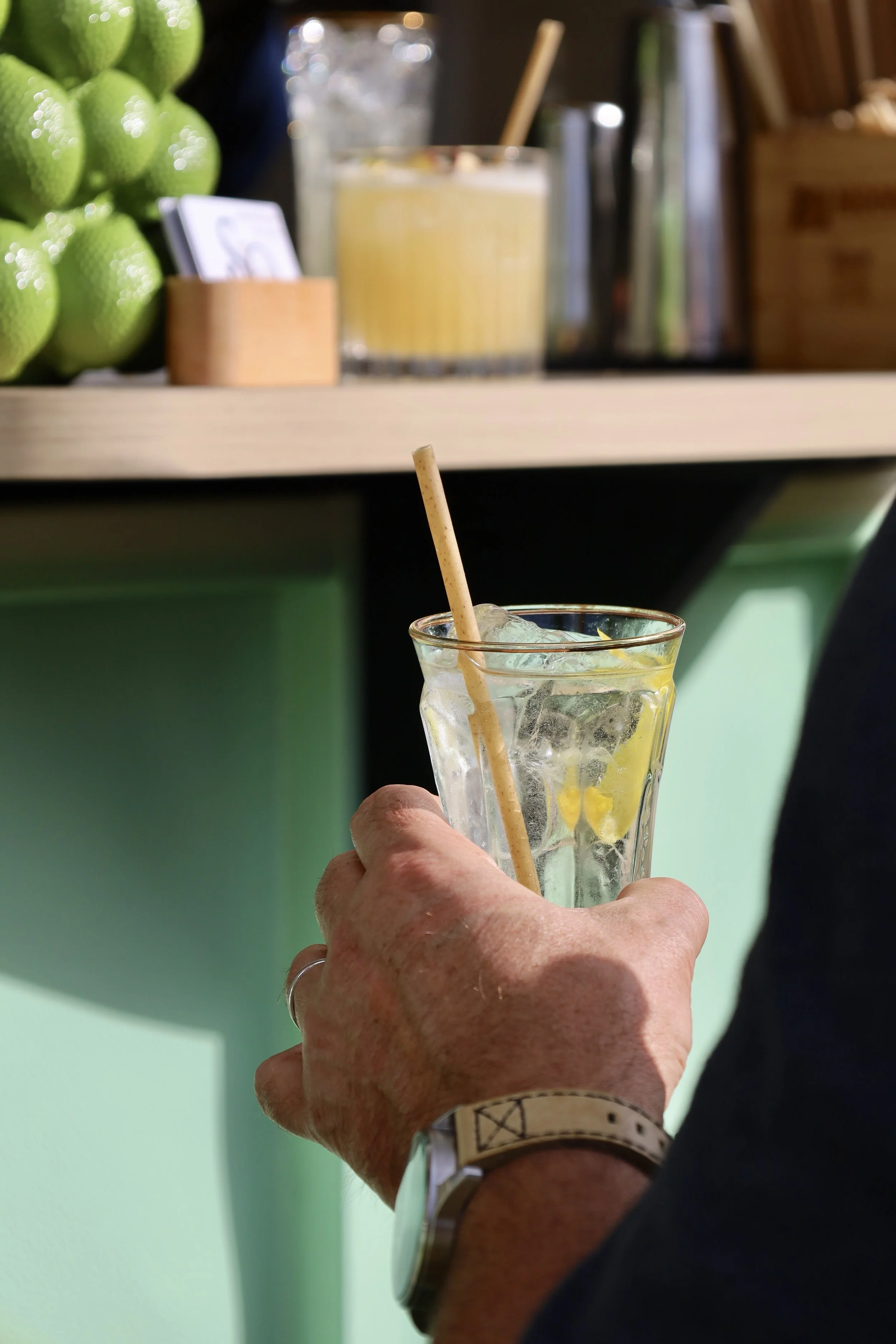Une main tenant un verre rempli d'eau avec glaçons, une tranche de citron et une paille en bois, dans un décor intérieur avec un meuble vert et plusieurs objets en arrière-plan.