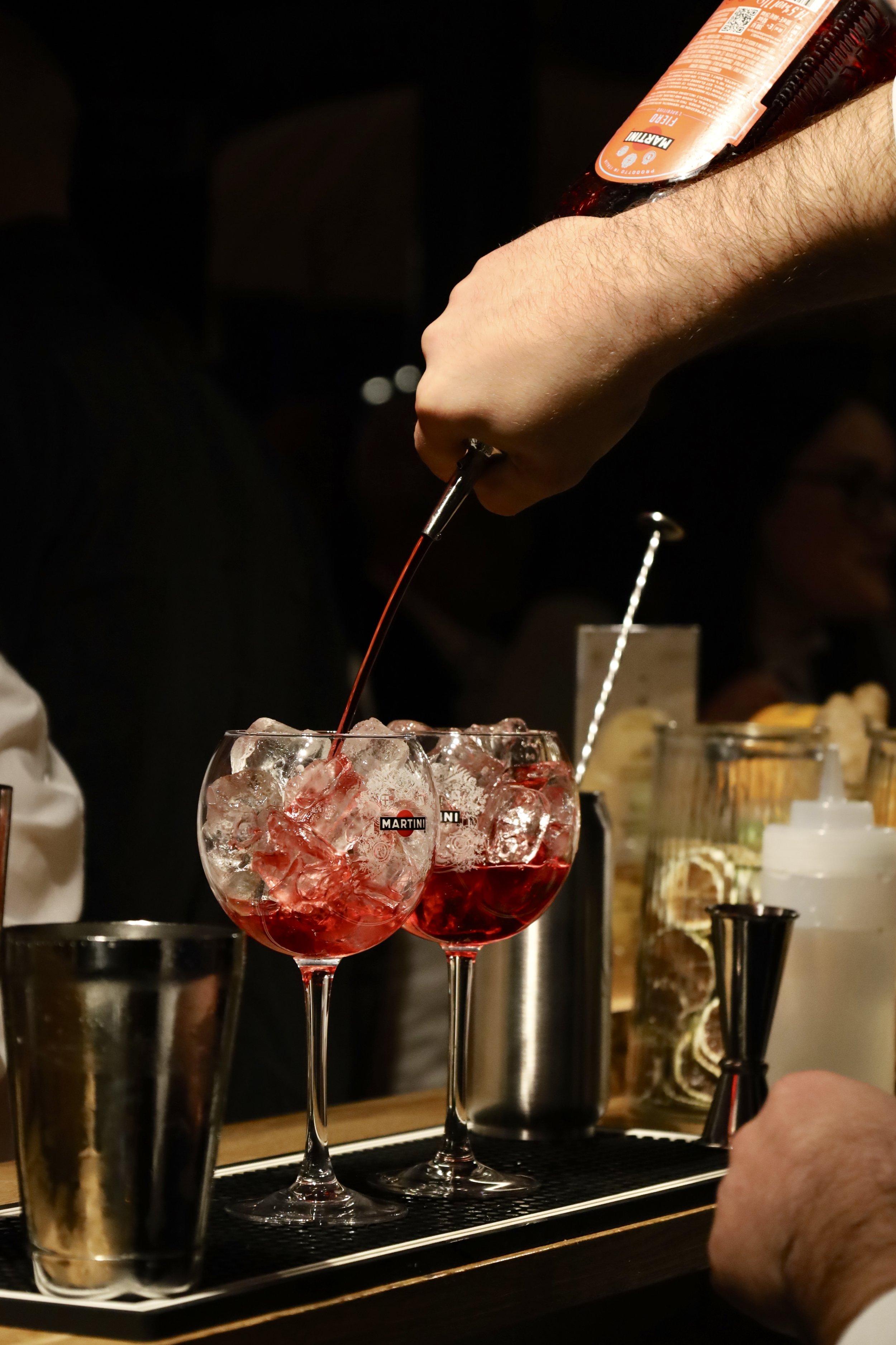 Un homme verse un cocktail rouge dans deux grands verres remplis de glaçons, au bar.
