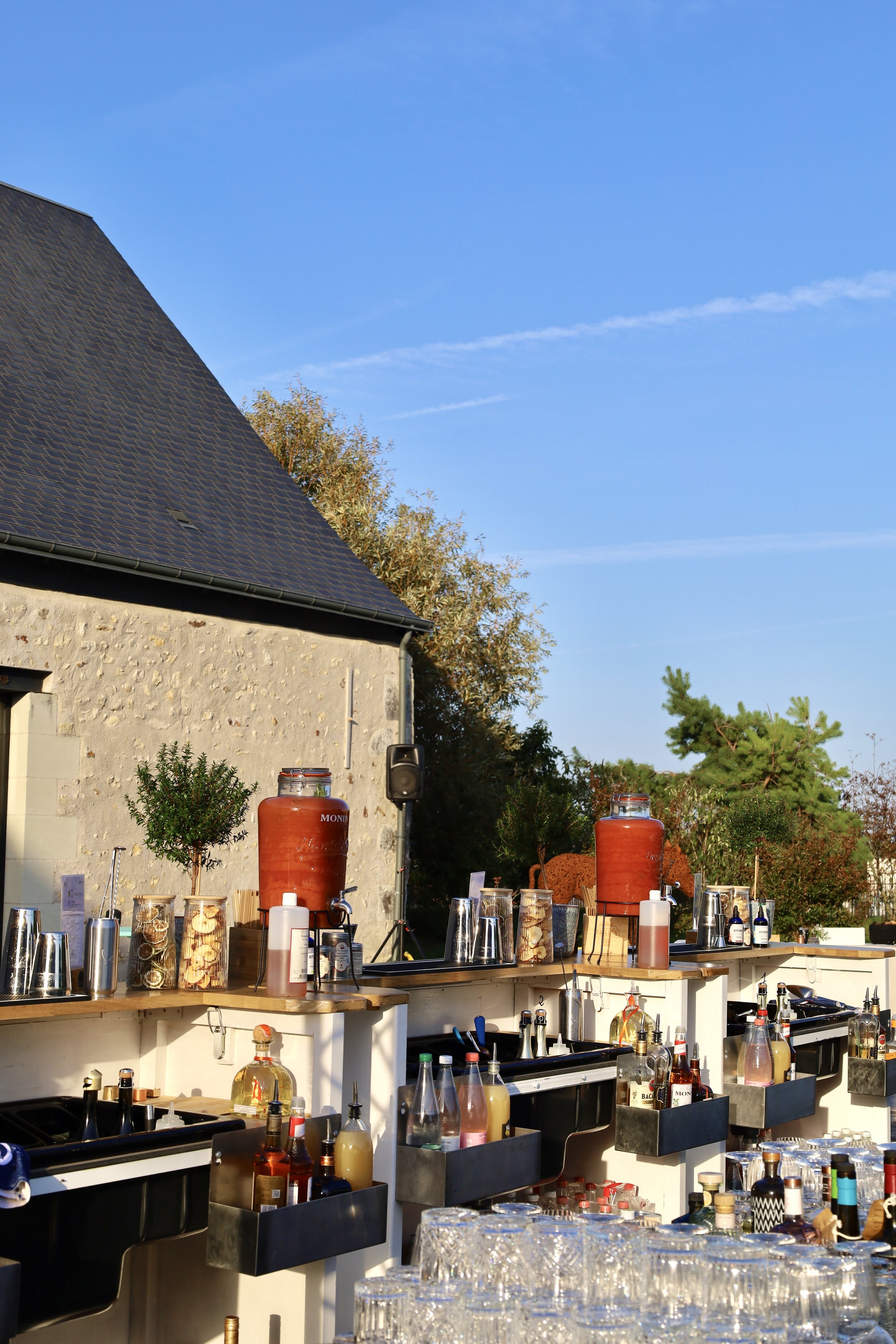 Bar extérieur en plein air avec des bouteilles, verres, et jarres de boisson, sous un ciel bleu, avec une maison en pierre en arrière-plan.