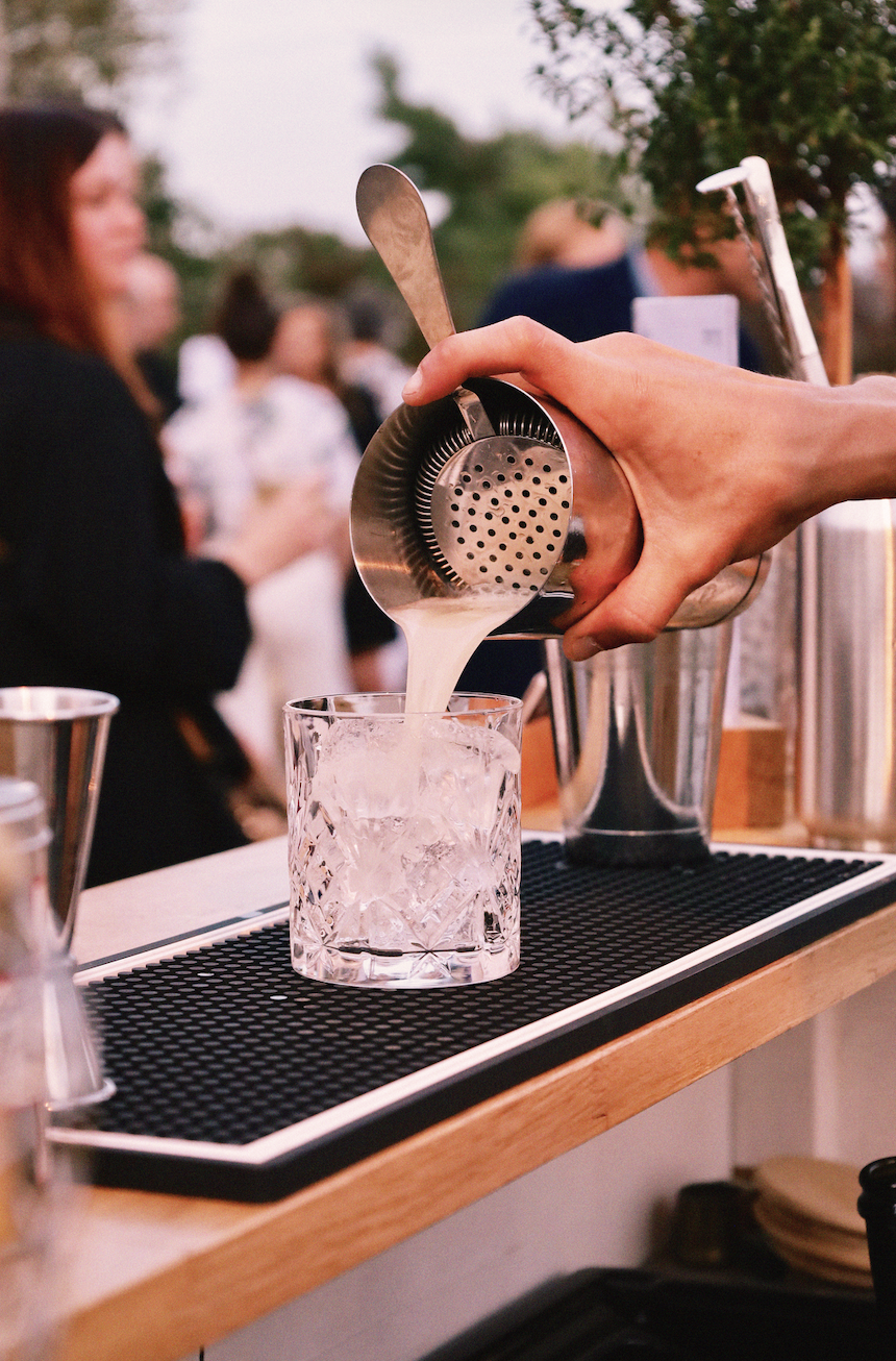 Une main verse un cocktail dans un verre rempli de glaçons lors d'une soirée en plein air, avec des personnes floues en arrière-plan.