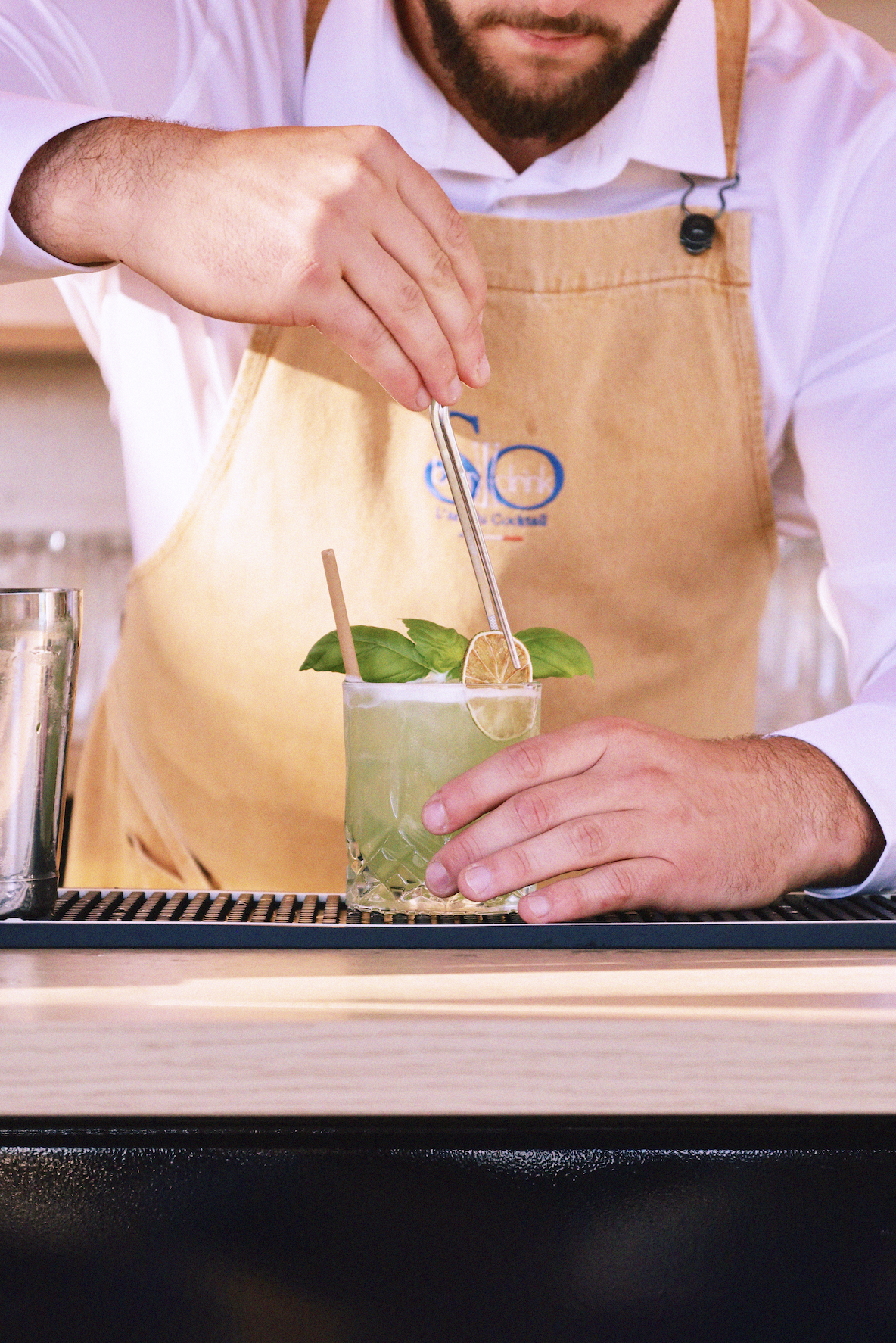 Un barman prépare un cocktail vert dans un verre, décoré avec des feuilles de basilic, un citron et une cannette, portant un tablier beige.