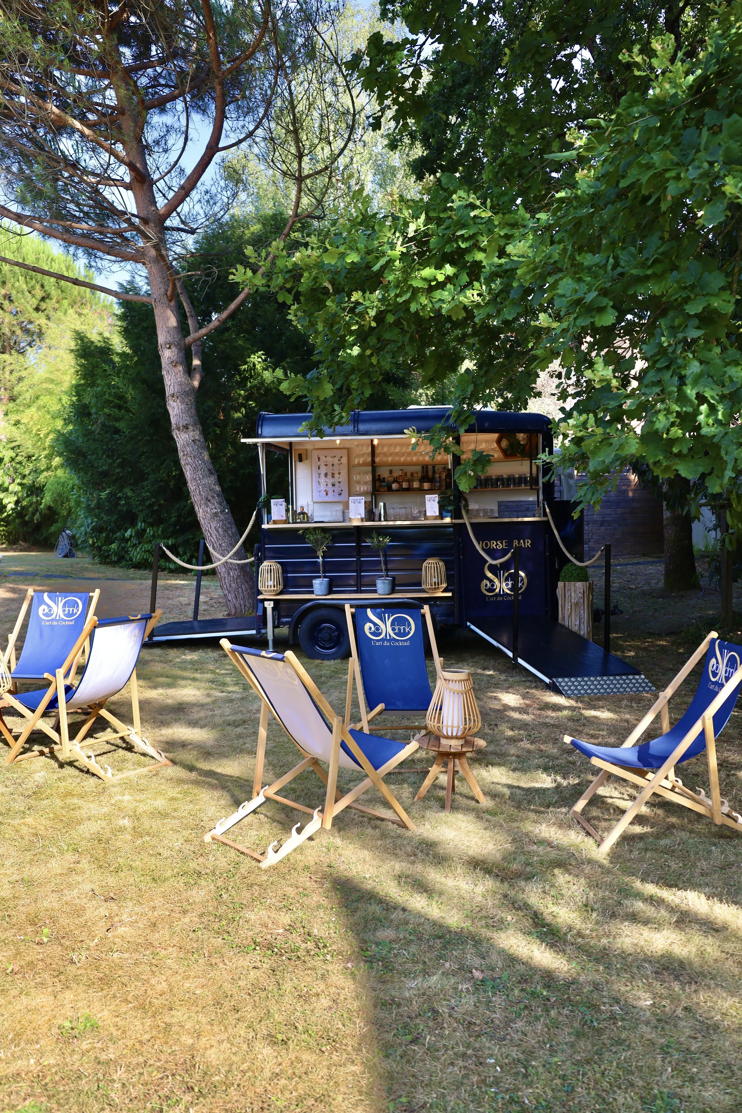 Un bar mobile en plein air avec chaises en bois et toiles bleues, entouré d'arbres verts sous un ciel ensoleillé.