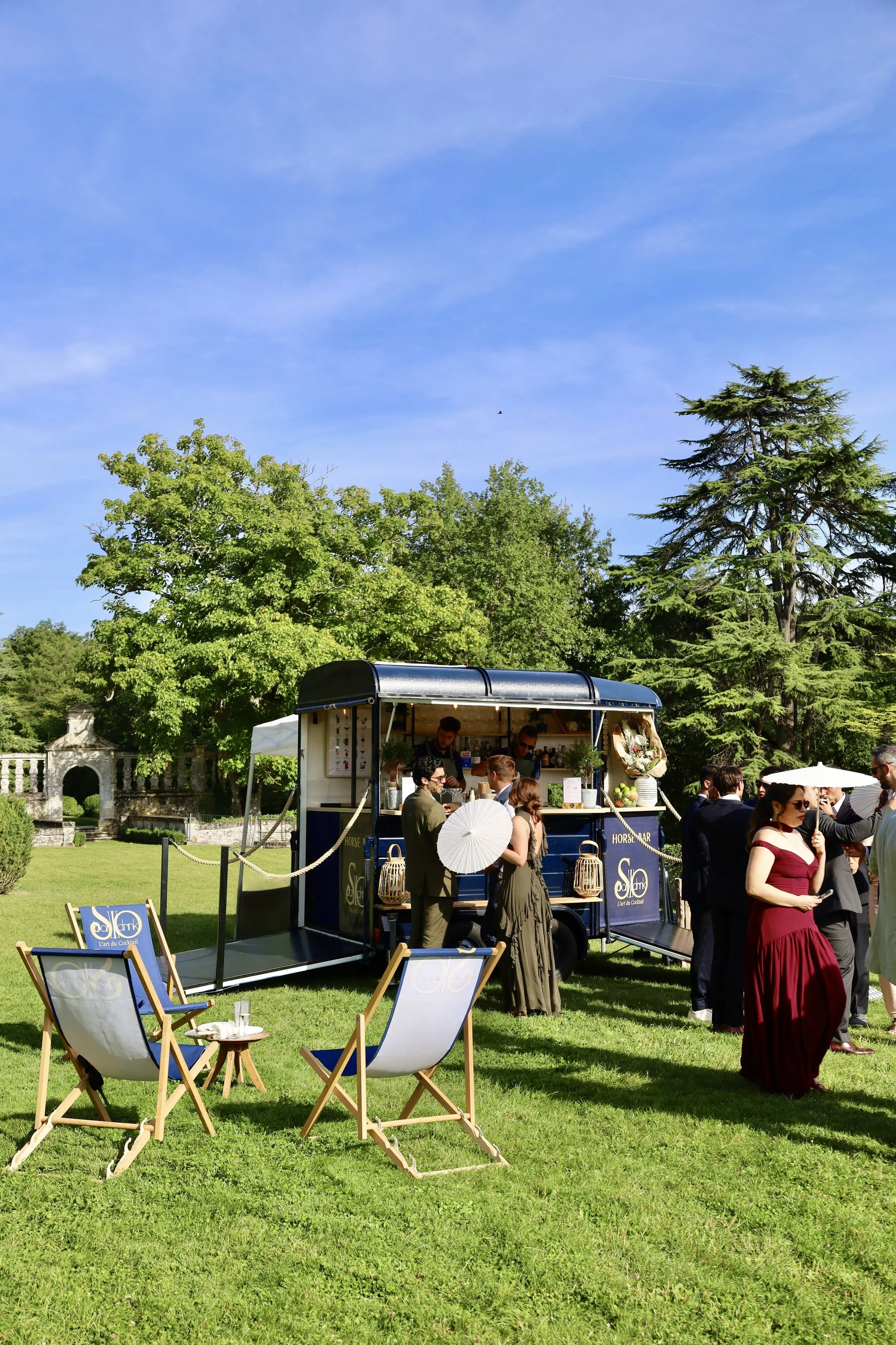 Un groupe de personnes habillées élégamment dans un jardin avec un traiteur mobile, sous un ciel bleu.