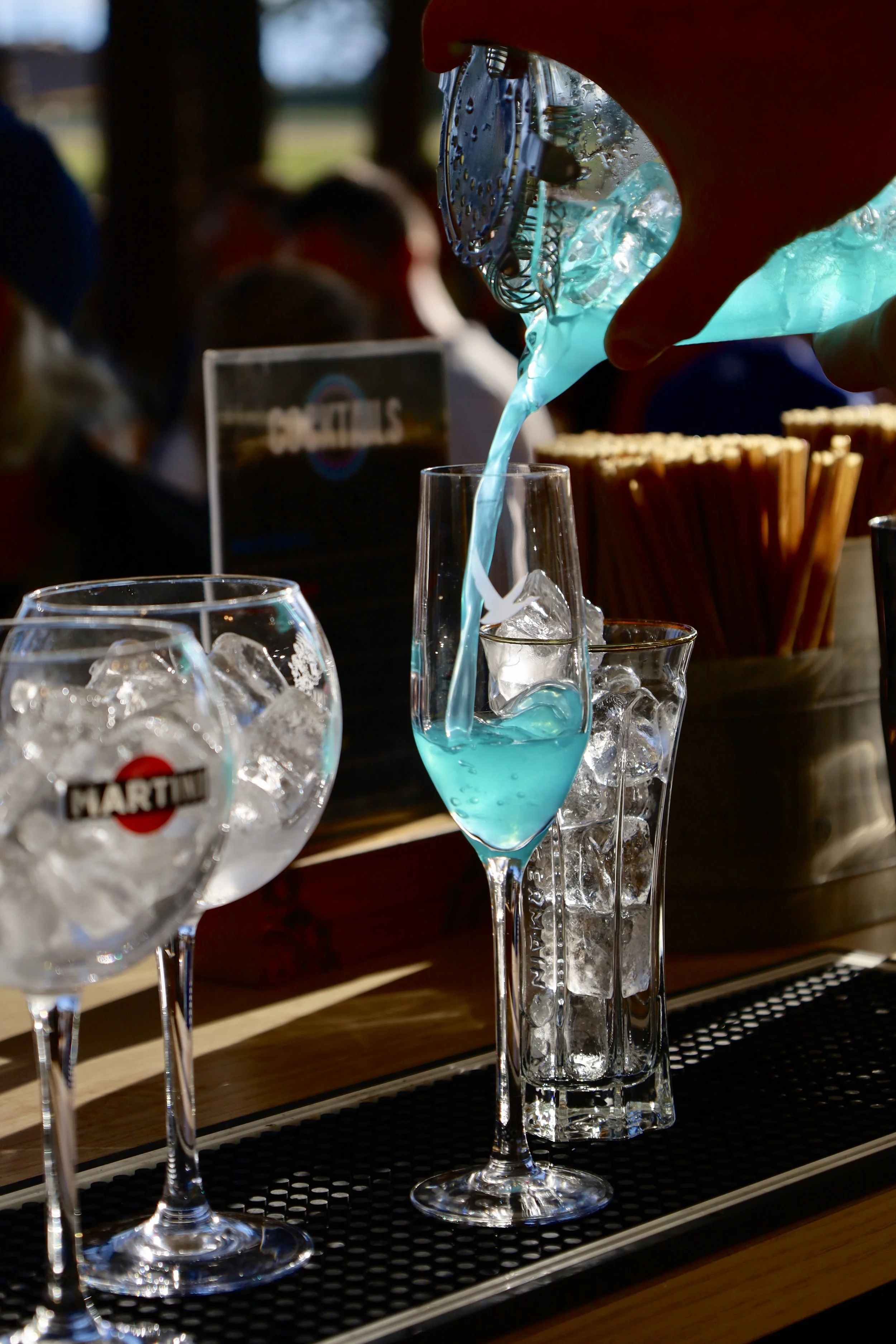 Une personne verse une boisson bleue dans une flûte à champagne dans un bar. Plusieurs verres avec des glaçons et une bouteille d'eau gazeuse sont visibles, ainsi qu'une panière de spaghetti en arrière-plan.