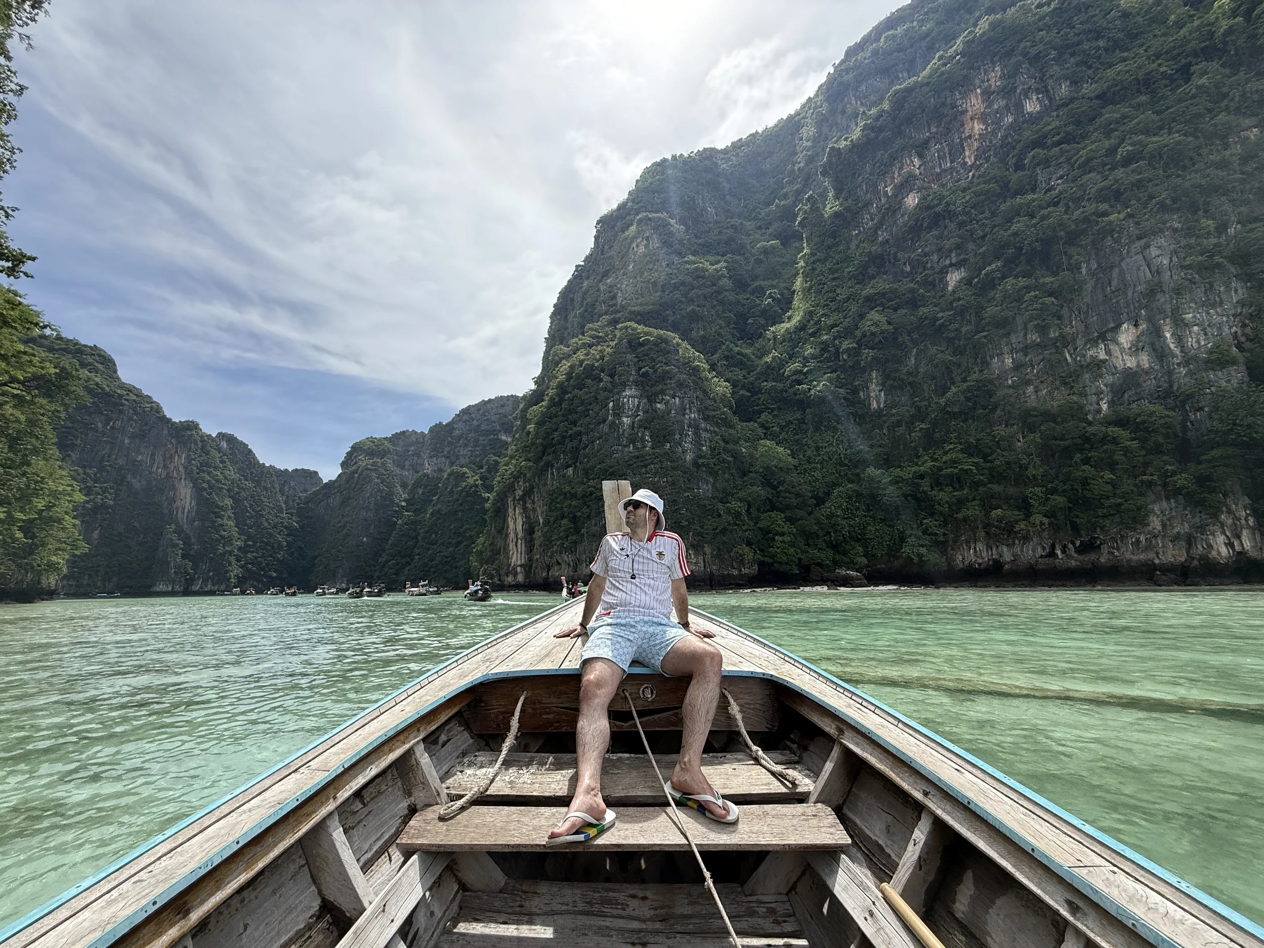 A person sitting at the front of a wooden boat on a turquoise waterway, with large limestone cliffs covered in green vegetation on either side under a cloudy sky.
