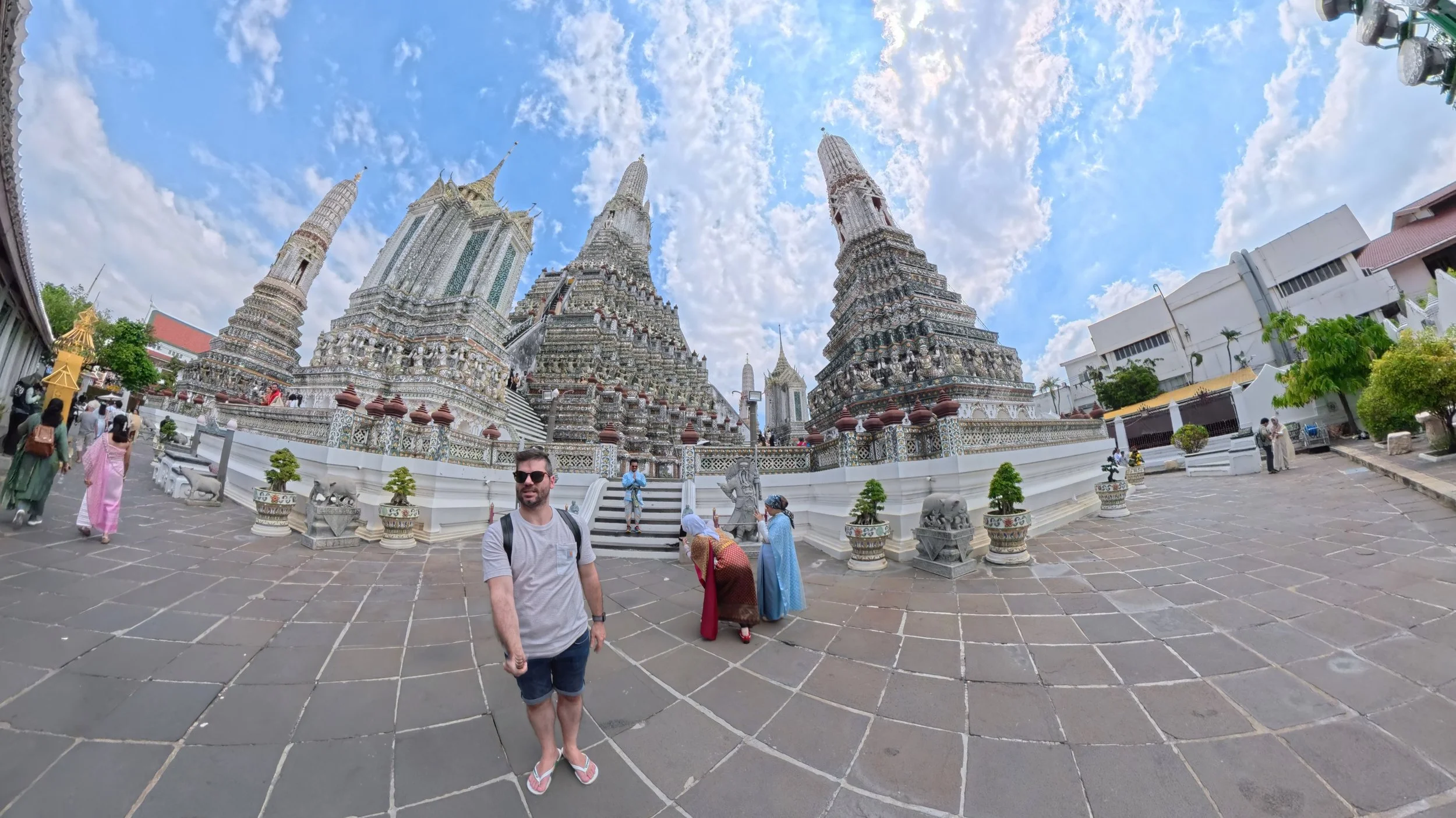 Tourists visiting the Wat Arun temple in Bangkok, Thailand, with ornate spires and intricate detailing under a partly cloudy sky.