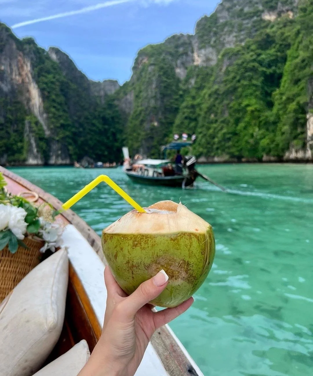 A person holding a coconut with a yellow straw on a boat in a tropical waterway surrounded by tall green cliffs and boats in the distance.