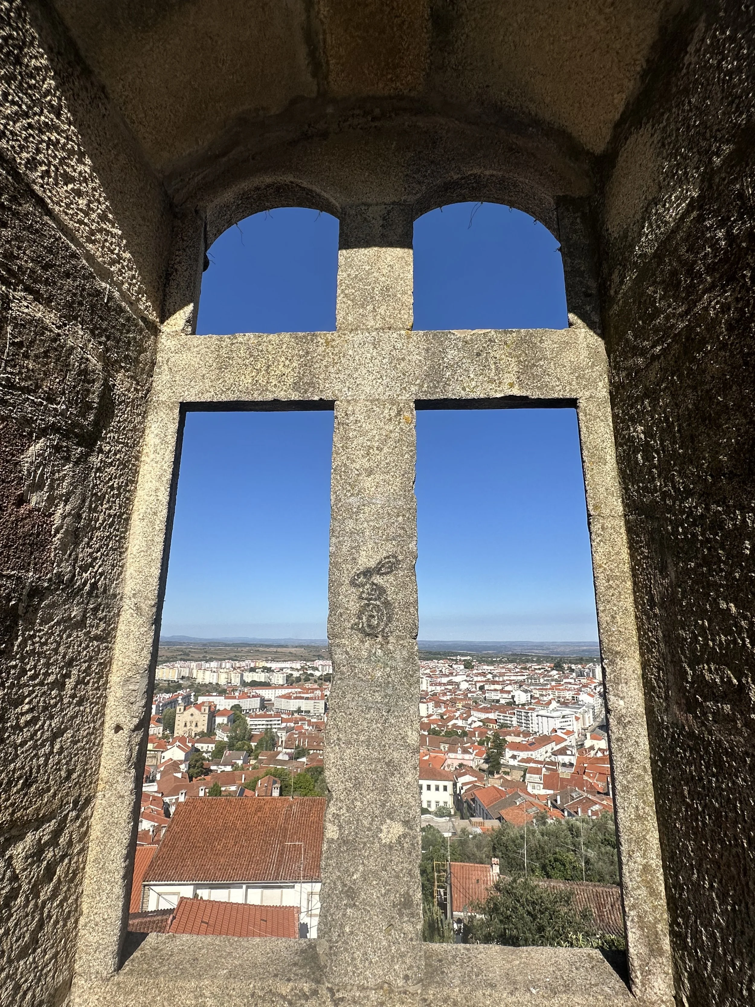 View through a stone cross-shaped window showing a city with red rooftops and a clear blue sky.