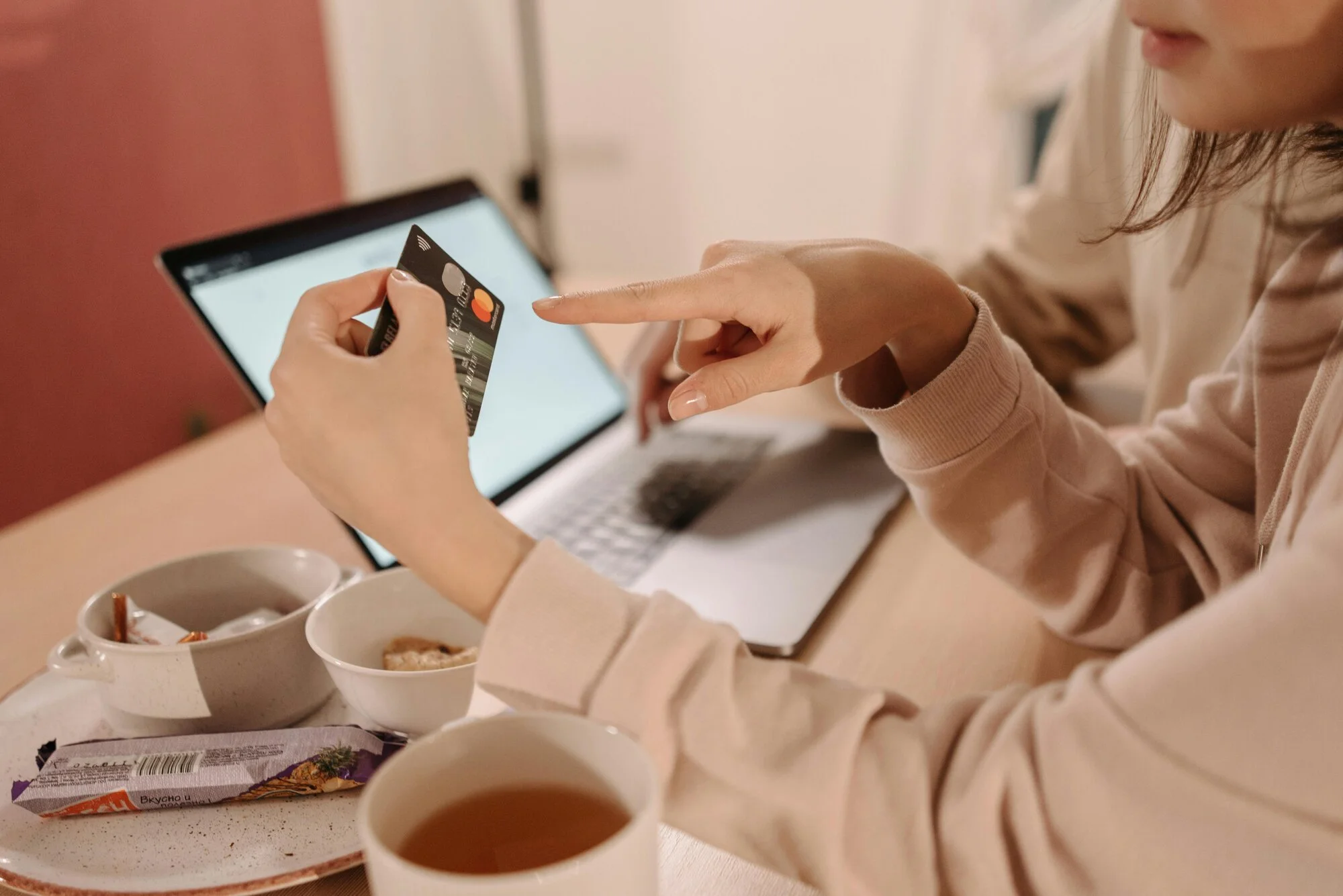 A woman is holding a black credit card while shopping online on her laptop, with a cup of tea and snacks on the table.