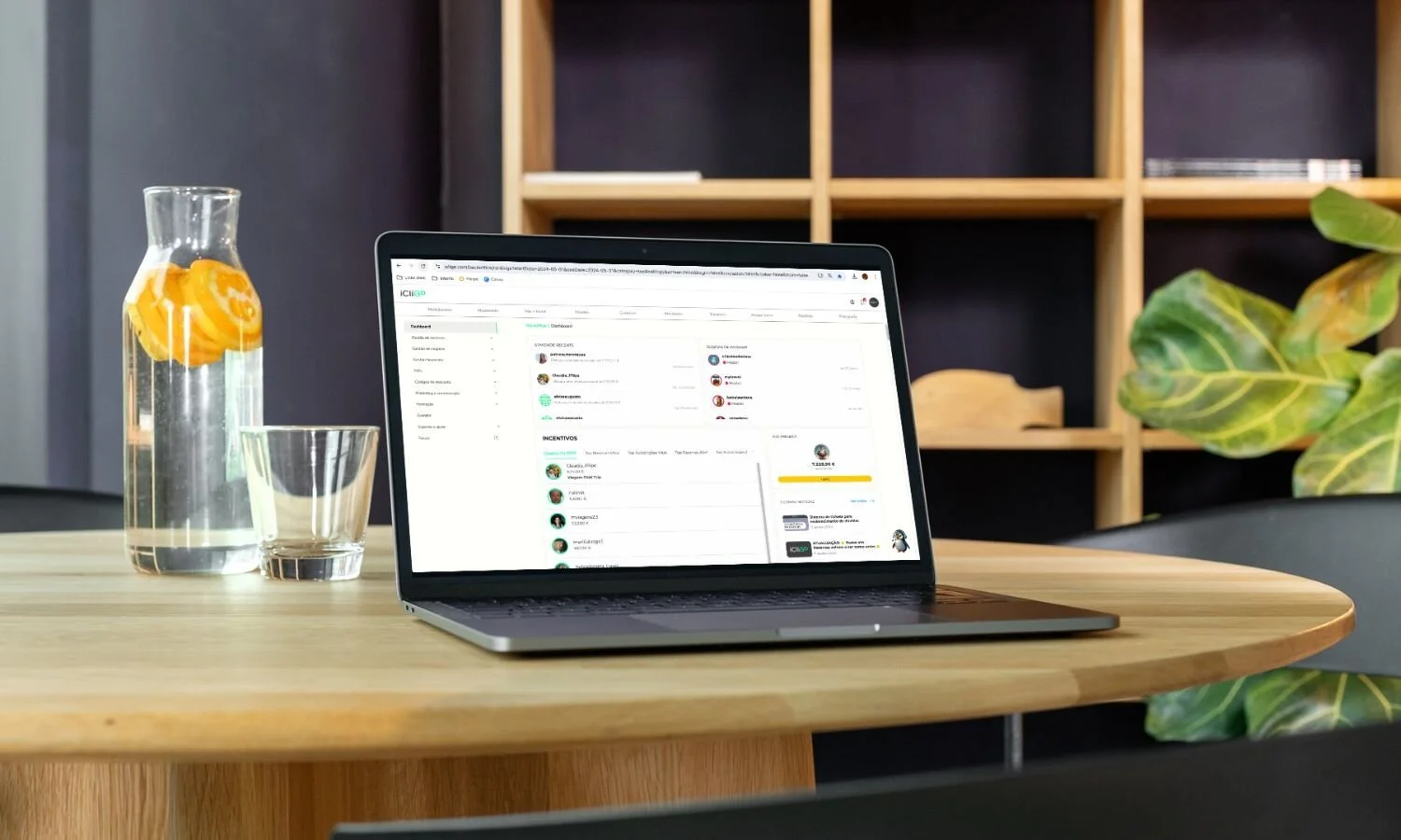 Laptop on a wooden table displaying a dashboard, with a glass pitcher of water with lemon slices, an empty glass, and a plant nearby in a modern office or home setting.