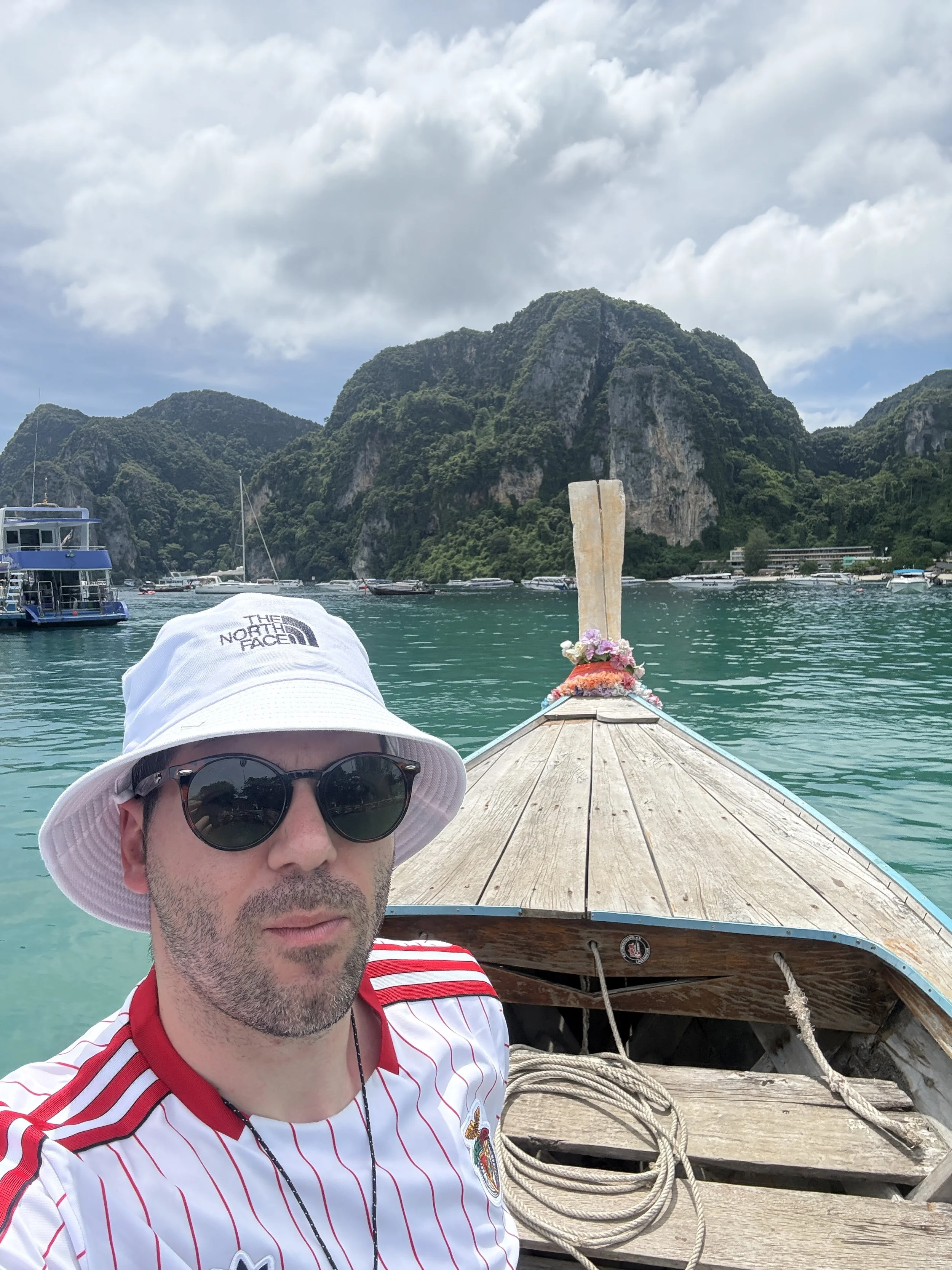 A man wearing a white bucket hat and sunglasses taking a selfie on a boat with a scenic tropical island and turquoise water in the background.