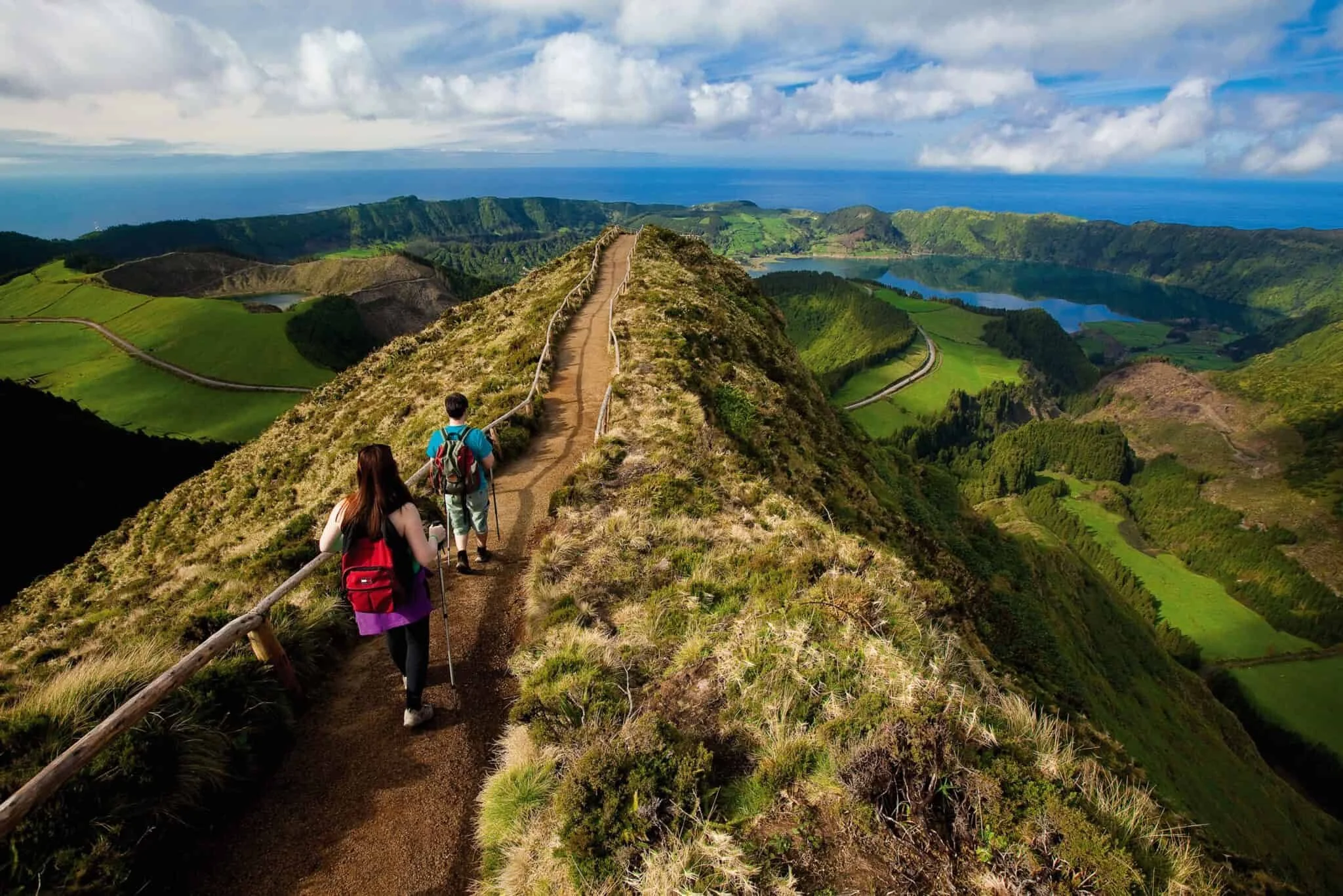 Three hikers walking along a narrow path on a mountain ridge with green rolling hills, lakes, and the ocean in the background under partly cloudy skies.