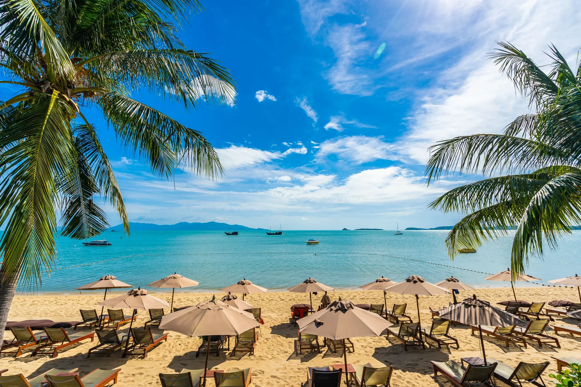 Sandy beach with sun loungers and umbrellas under palm trees, calm turquoise water with boats, blue sky with clouds, distant islands.
