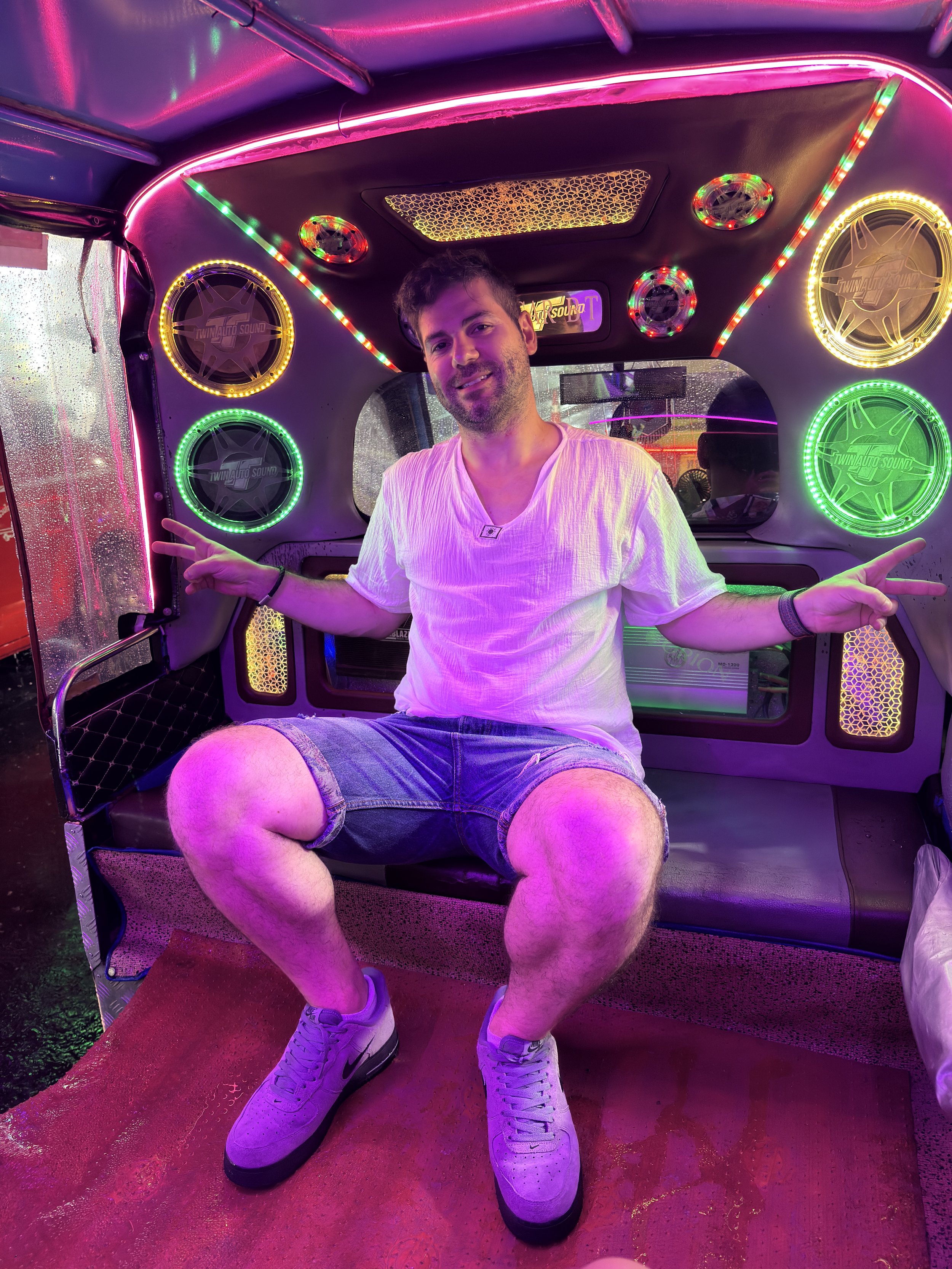 A man sitting inside a neon-lit tuk-tuk, smiling and making peace signs with both hands. The tuk-tuk is decorated with colorful LED lights and speakers.