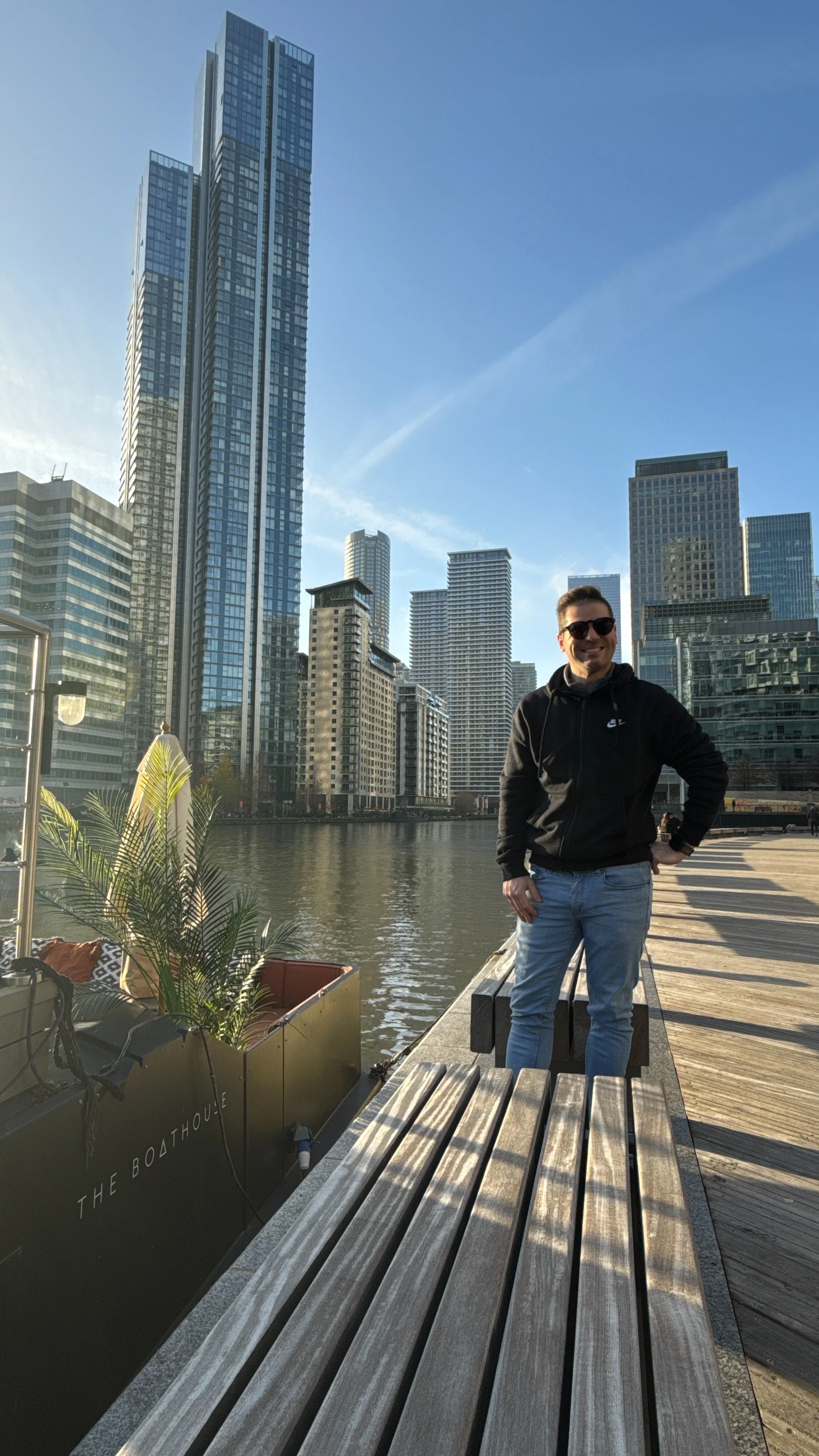 A man standing on a wooden dock by the water with a city skyline featuring tall skyscrapers behind him, wearing sunglasses, a black hoodie, and jeans, smiling at the camera.