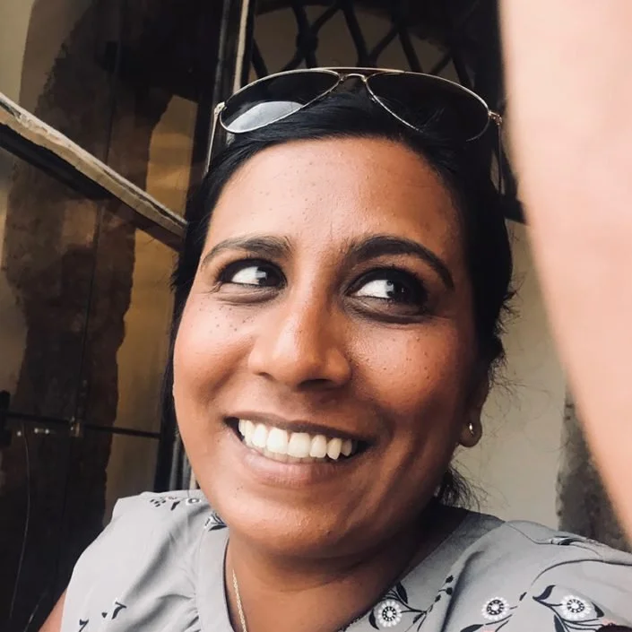 Close-up of a woman smiling, wearing sunglasses on her head and a light-colored top, with dark hair tied back.