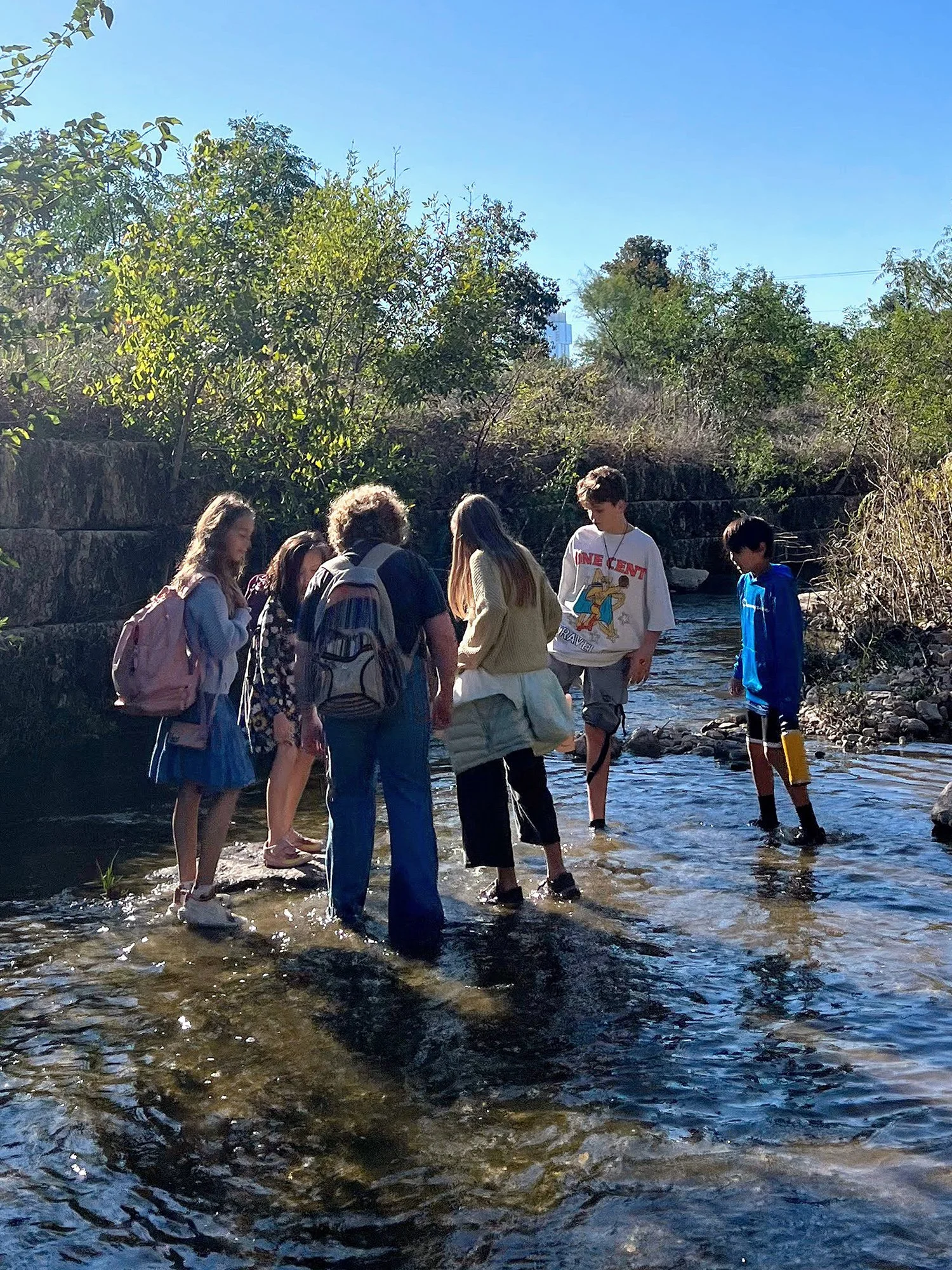 Group of children with backpacks wading in a shallow creek surrounded by greenery on a sunny day.