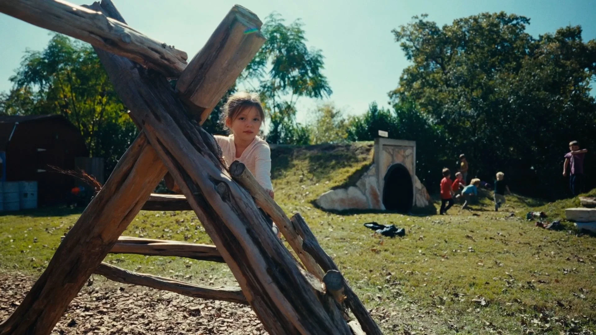 A young girl peering from behind a wooden climbing frame outdoors on a sunny day, with other children playing in the background near a tunnel structure and trees.