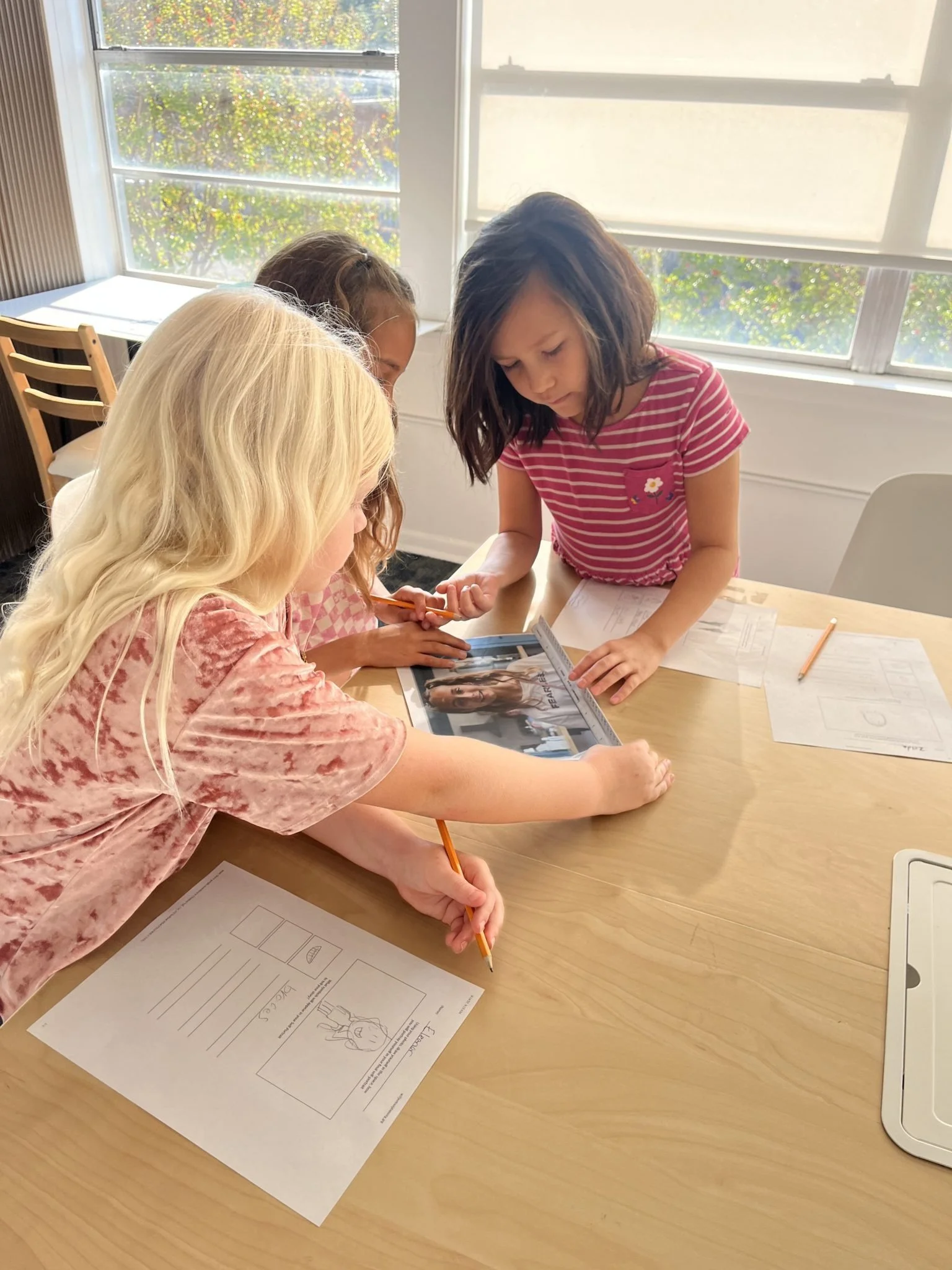 Four young girls gathered around a table, examining a magazine, with papers and pencils, in a bright room with large windows.
