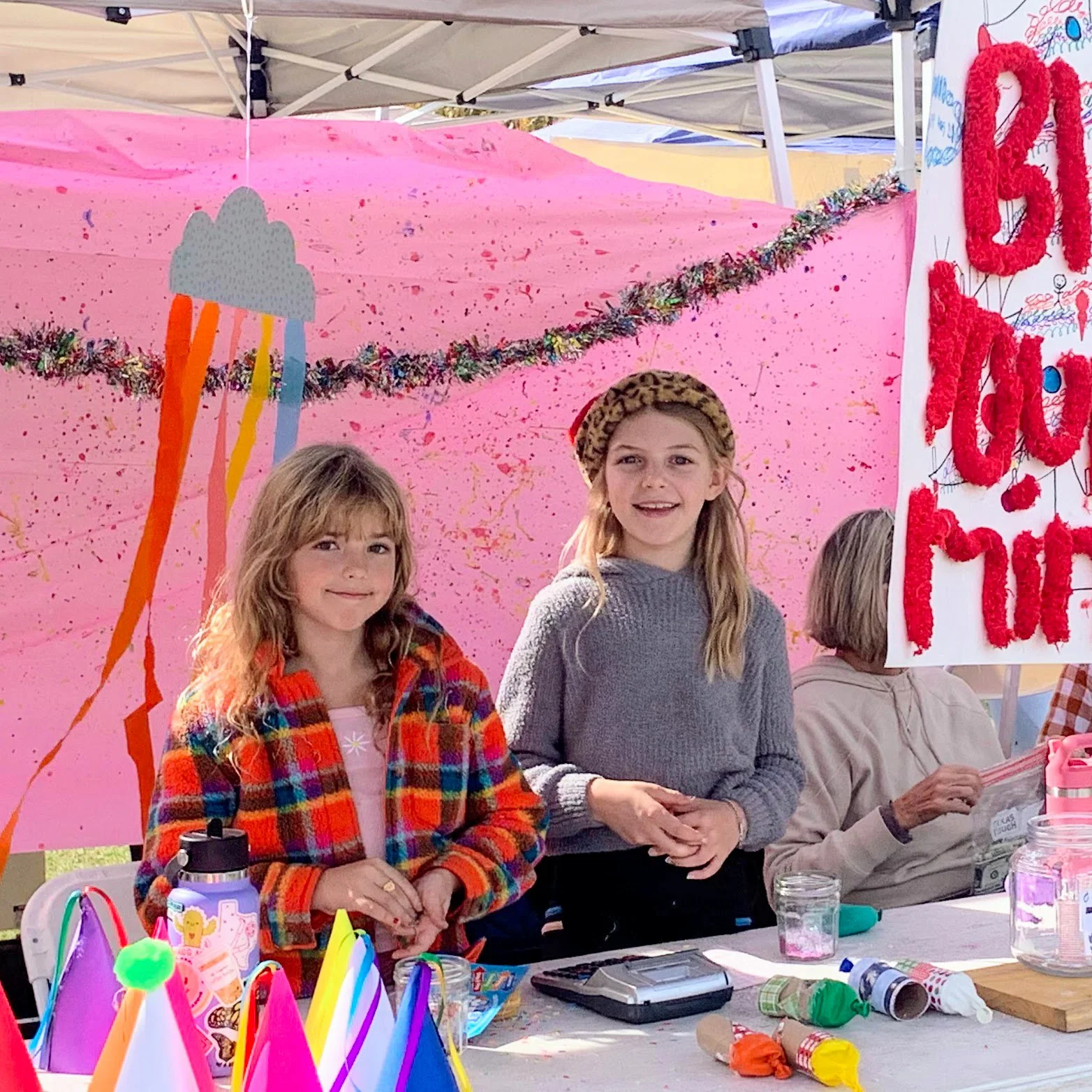 Two young girls at a birthday party, one wearing a colorful plaid jacket and the other a gray sweater with a leopard print beret, standing in front of a pink decorated backdrop with an ice cream themed banner.