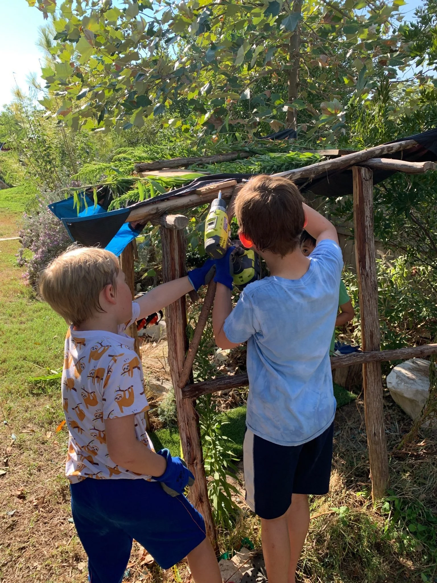 Three children working together on a gardening project, using a drill, under a small wooden and tarp shelter in a lush garden.