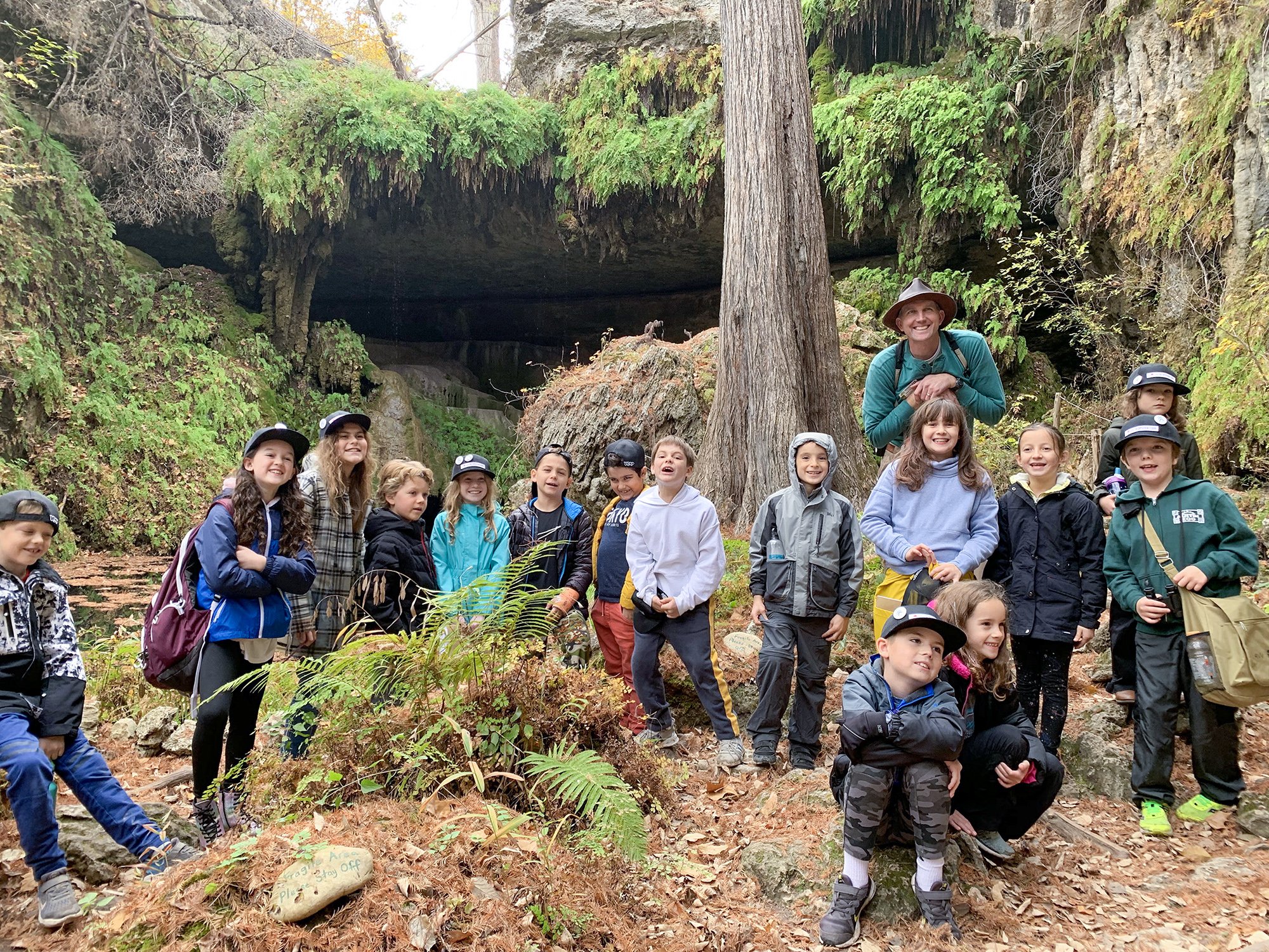 Group of children and their adult tour guide on a forest hiking trail near a rocky cave entrance, surrounded by trees and autumn foliage.